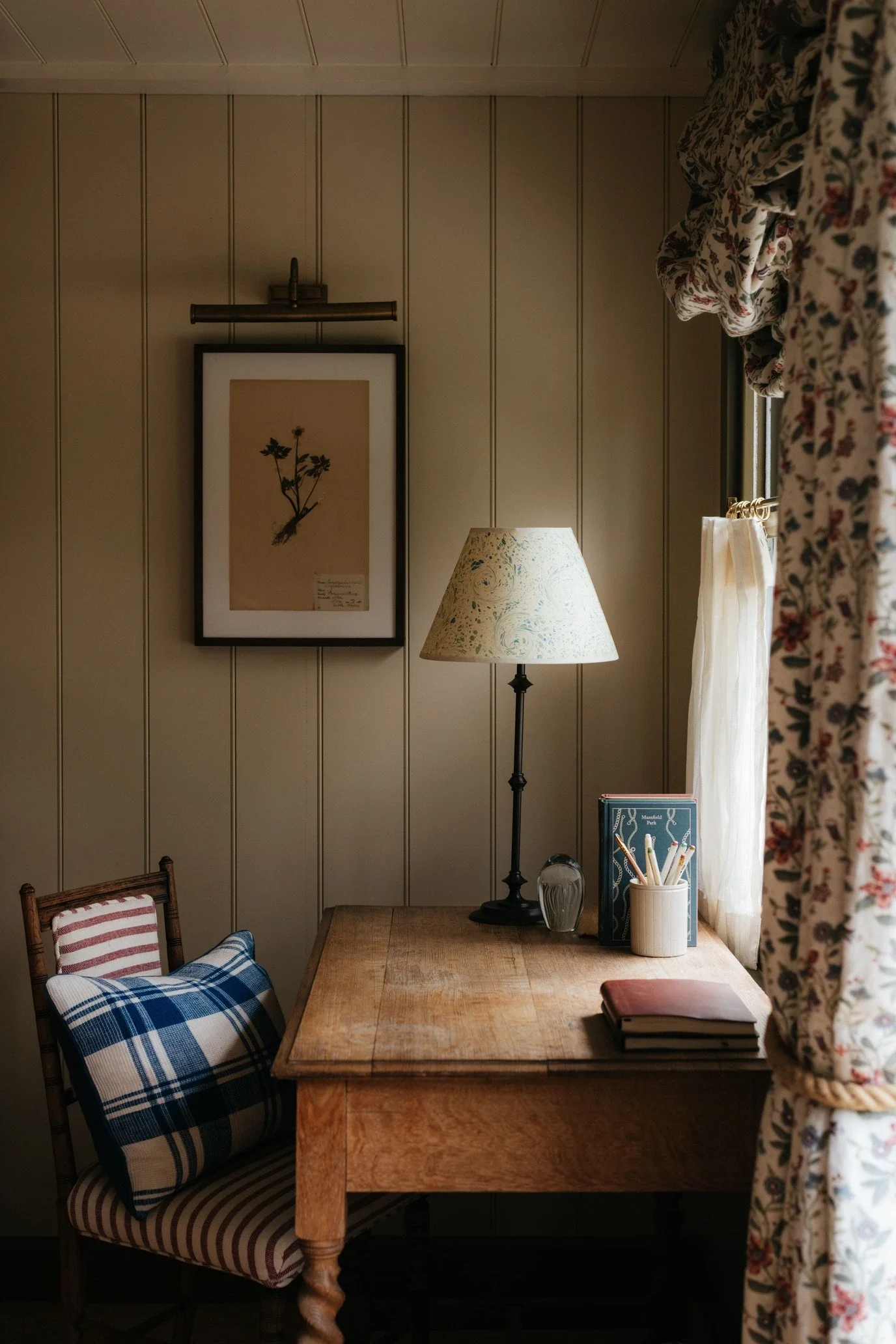 A cozy corner of a room featuring a wooden table, a plaid cushion on a wooden chair, and a table lamp with a floral lampshade. There are books, a glass, and a container with pencils on the table. The wall has vertical paneling, a framed botanical pri