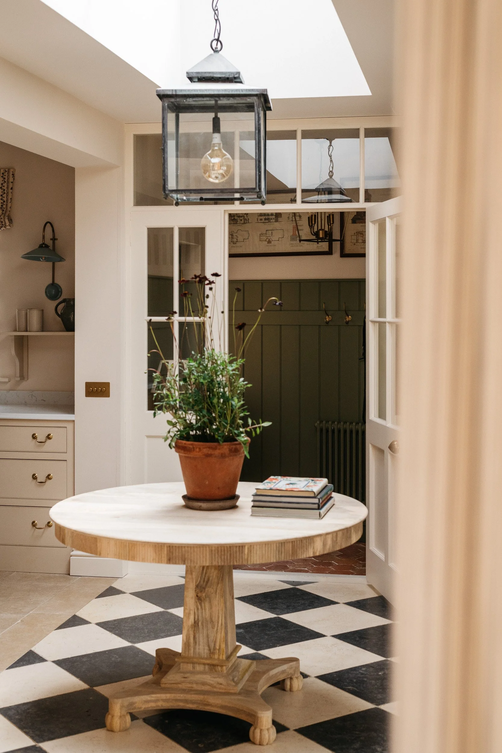 A round wooden table with a pottery vase containing a plant, and a stack of books on top. In the background, a kitchen with a dark green cabinet, framed artwork, and a white door opening into another room with checkered flooring and black-and-white t