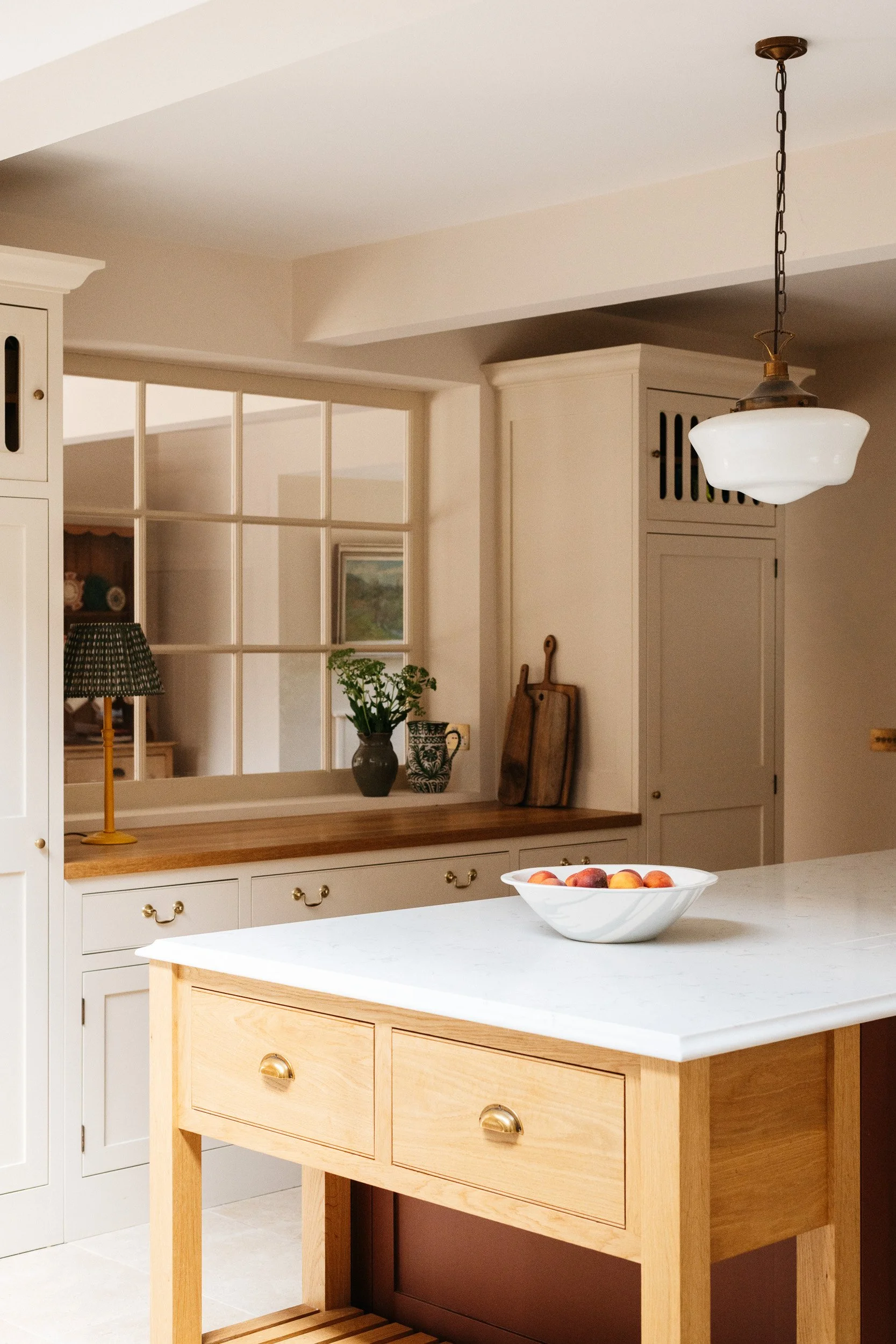 A kitchen island with a white marble top and light wood drawers. A bowl of peaches is on the island. In the background, there is a white cabinet, some decorative plates, a lamp, and a potted plant. A wooden cutting board and some other kitchen items 