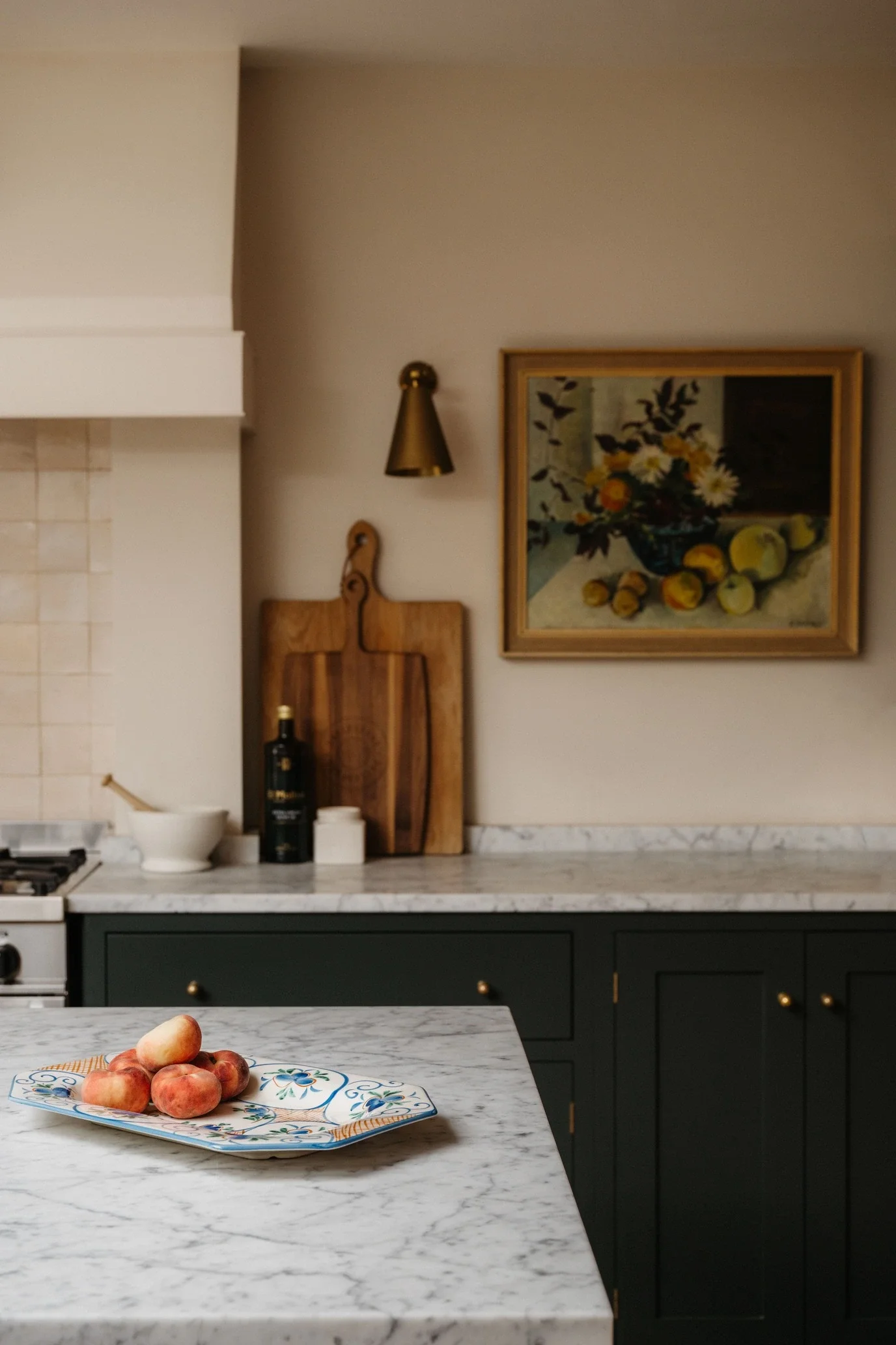 A kitchen with a white marble countertop, dark green cabinets, and a backsplash. On the counter, there is a plate with peaches, a wine bottle, and a bowl. In the background, there is a wooden cutting board, a framed painting of a flower vase with sun