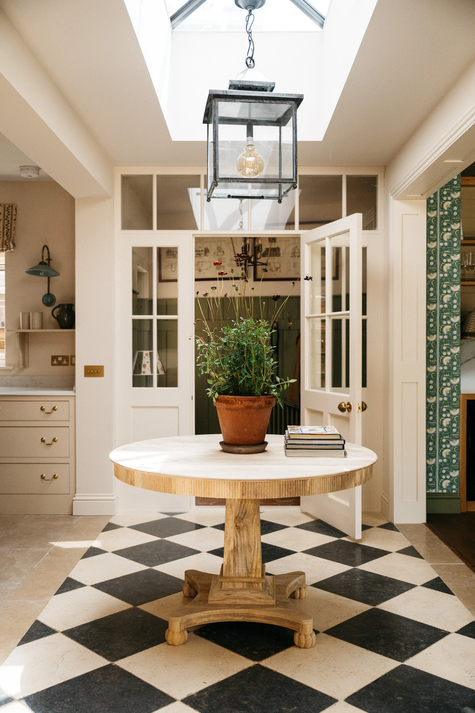 A kitchen or dining area with a round wooden table holding a potted plant and a stack of magazines, black and white checkered floor tiles, open white double doors with glass panes, and a skylight illuminating the space.