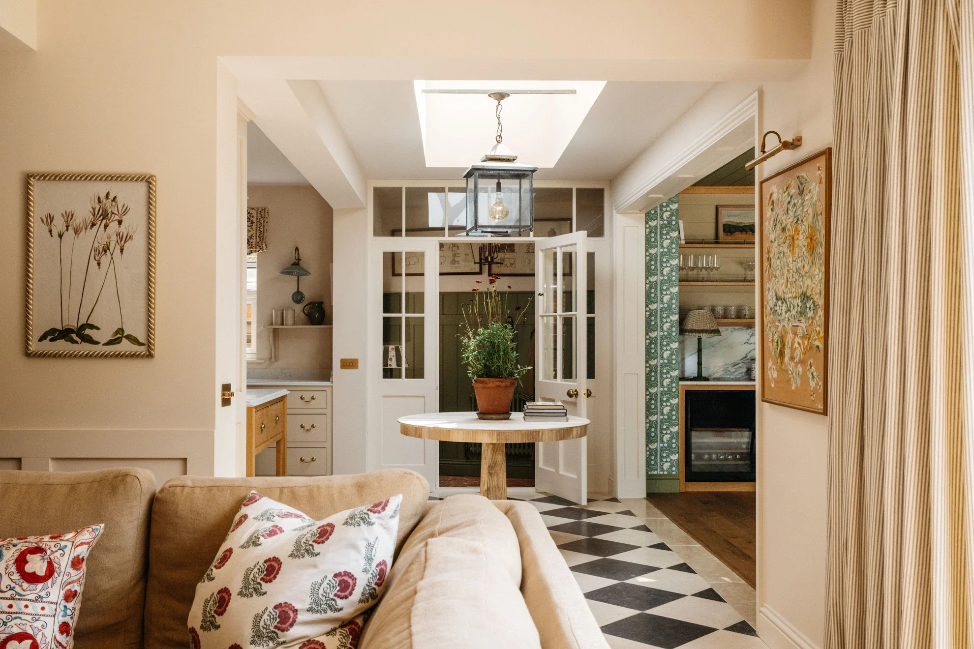 Living room with beige sofa, two decorative pillows, a round wooden table with a potted plant, open white double doors leading to another room, and artwork on the walls. Ceiling has a skylight with a hanging light fixture.