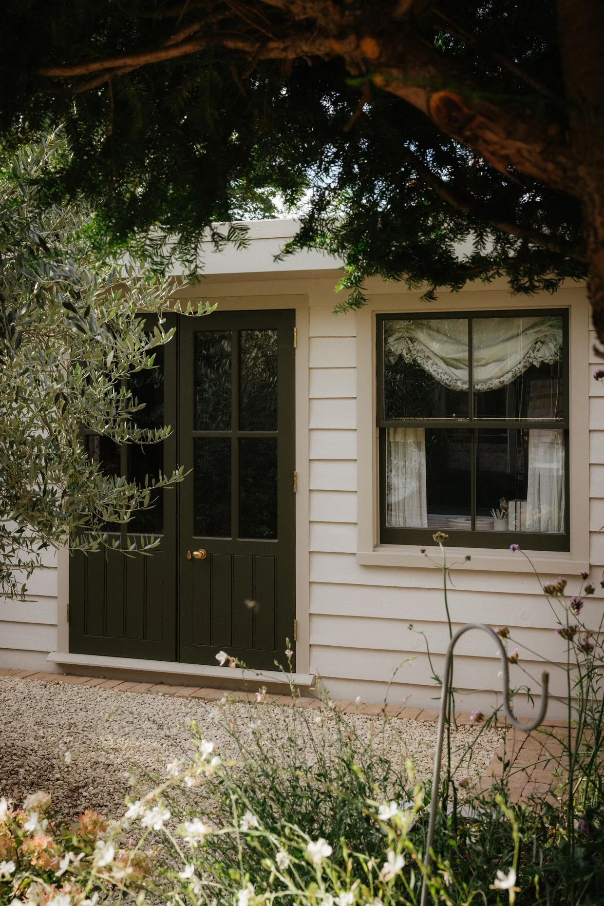 The photo shows the front of a house with a black door and a window, surrounded by greenery and flowering plants, taken during the daytime.