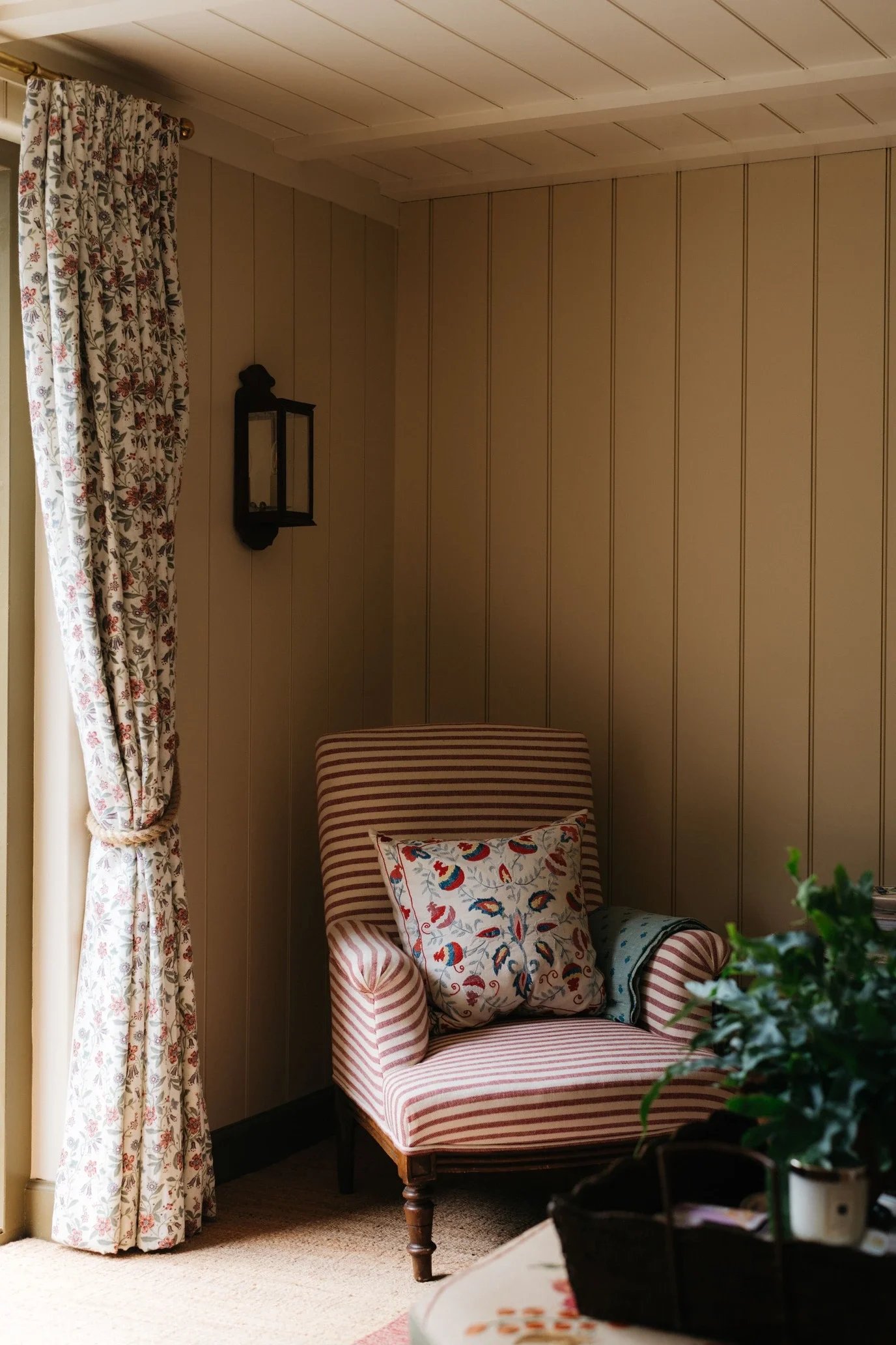 A cozy corner of a room with a vintage striped armchair, a decorative pillow with colorful feathers, a small side table with potted plants, beige wood-paneled walls, floral curtains, and a black wall-mounted lantern.