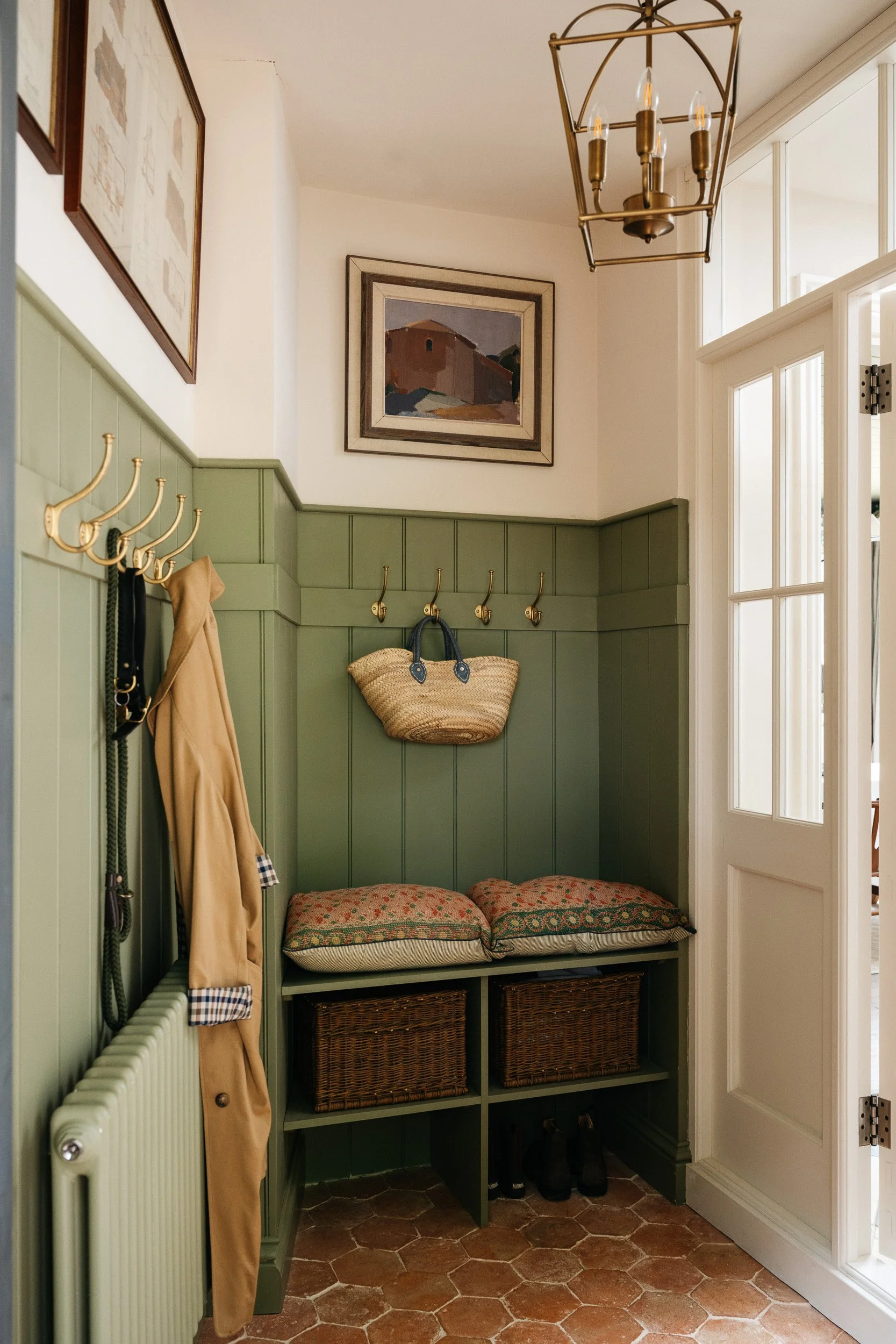 Entryway with green wainscoting, hooks with coats and bags, a wicker basket hanging on the wall, a bench with patterned cushions, wooden shelves with baskets underneath, a chandelier, and a white door with glass panels.
