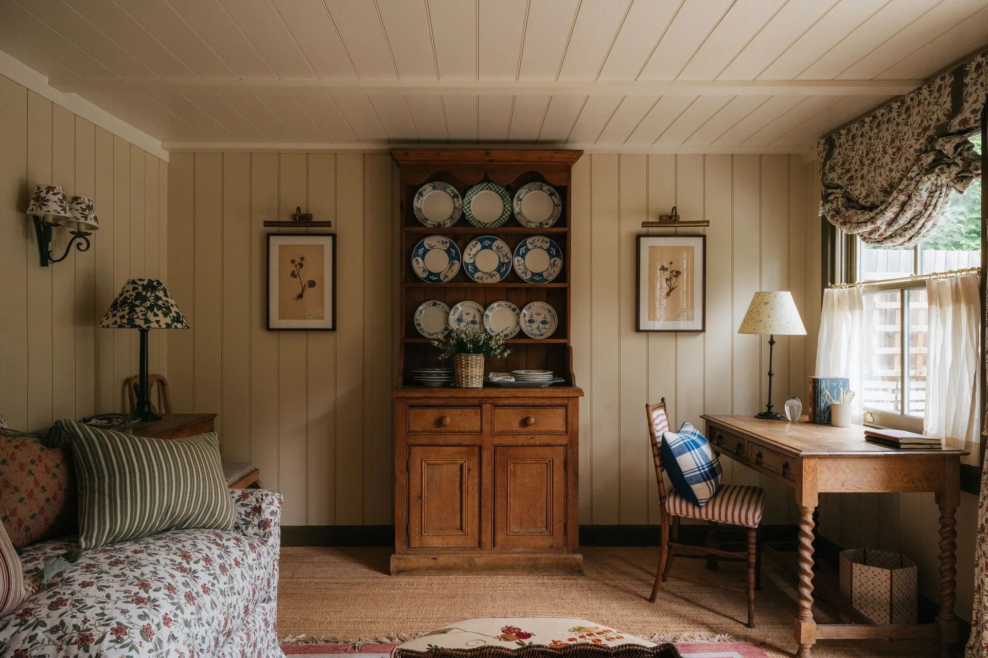 A cozy living room with wood-paneled walls, a floral-patterned sofa, a wooden cabinet displaying decorative plates, framed botanical artwork, a desk near window with a striped chair, and floral curtains pulled to the side.