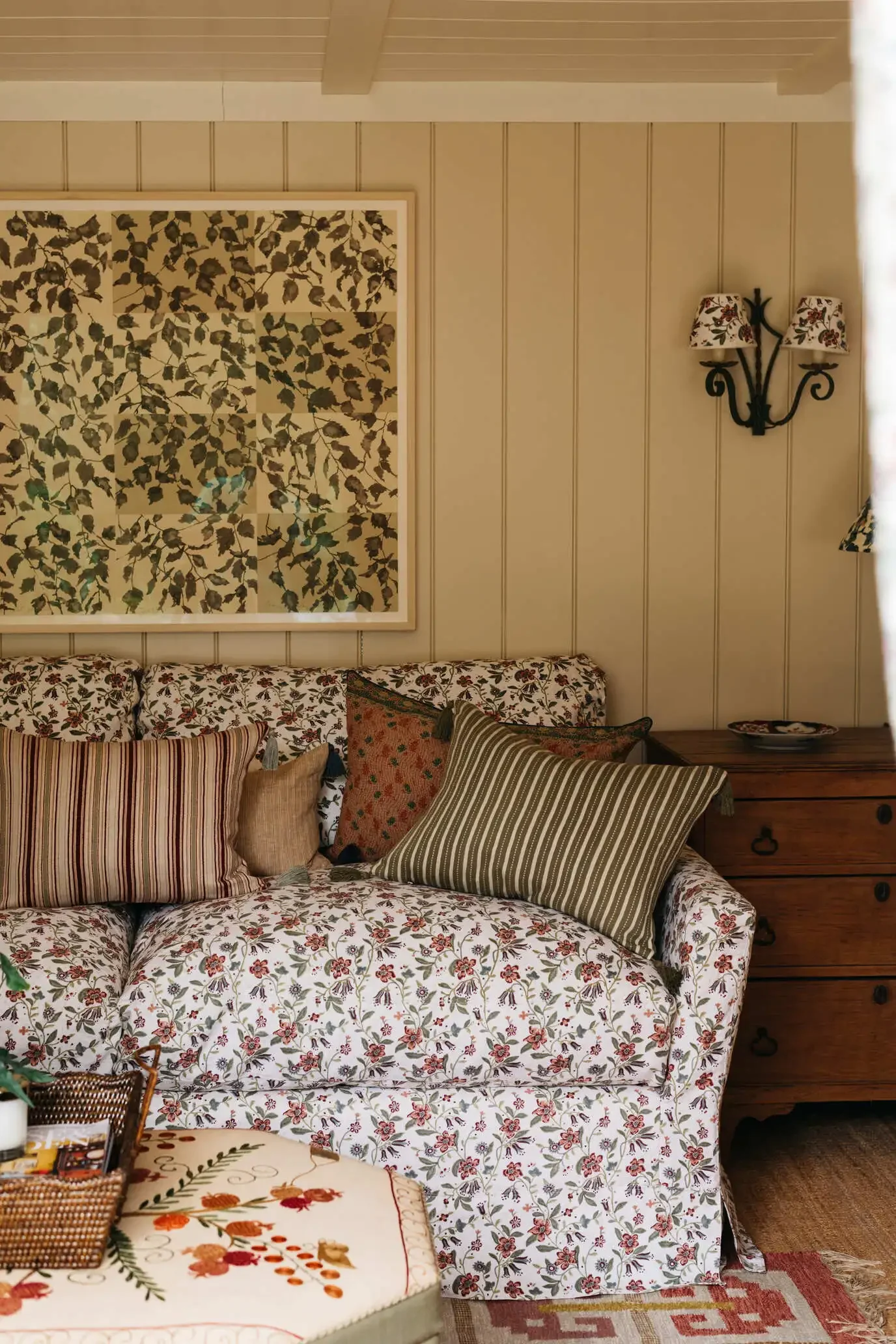 A living room corner with a floral-patterned sofa, several decorative pillows, a wooden side table, a wall-mounted lamp with floral lampshades, beige paneled walls, and a large framed artwork of leaf patterns.