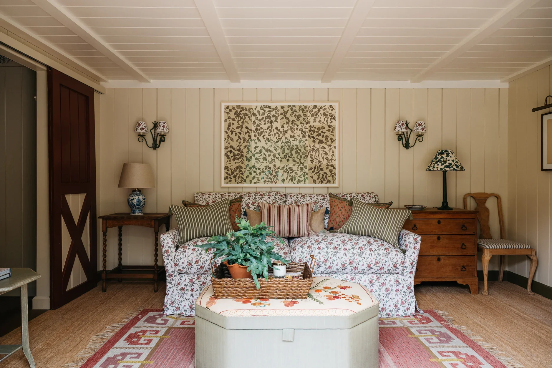 Living room with floral sofa, various decorative pillows, a wooden side table with a lamp, wall art, and a plant