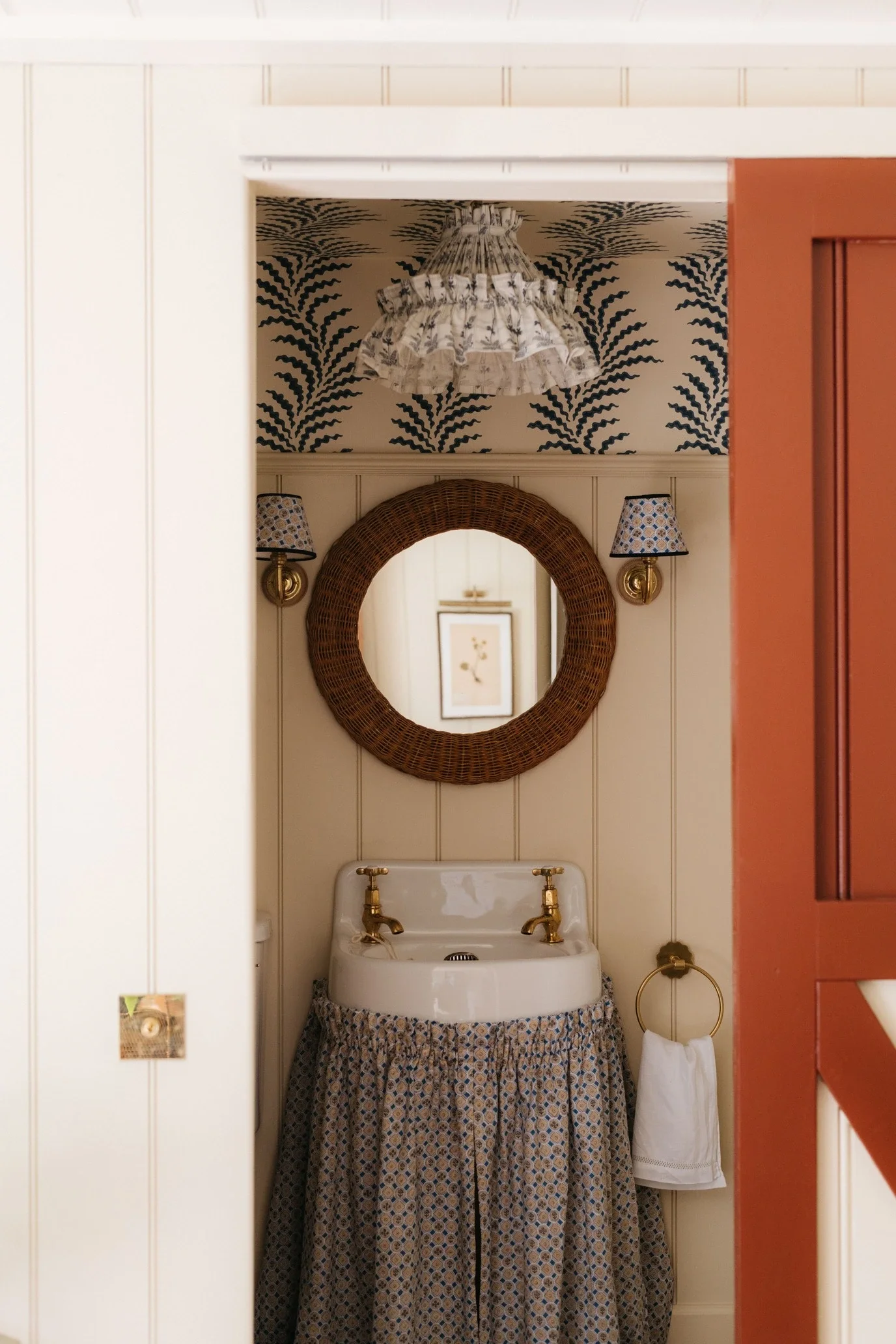 Interior view of a vintage bathroom with a white sink, gold fixtures, a round wicker mirror, nautical themed wallpaper, and a decorative fabric curtain covering the sink.