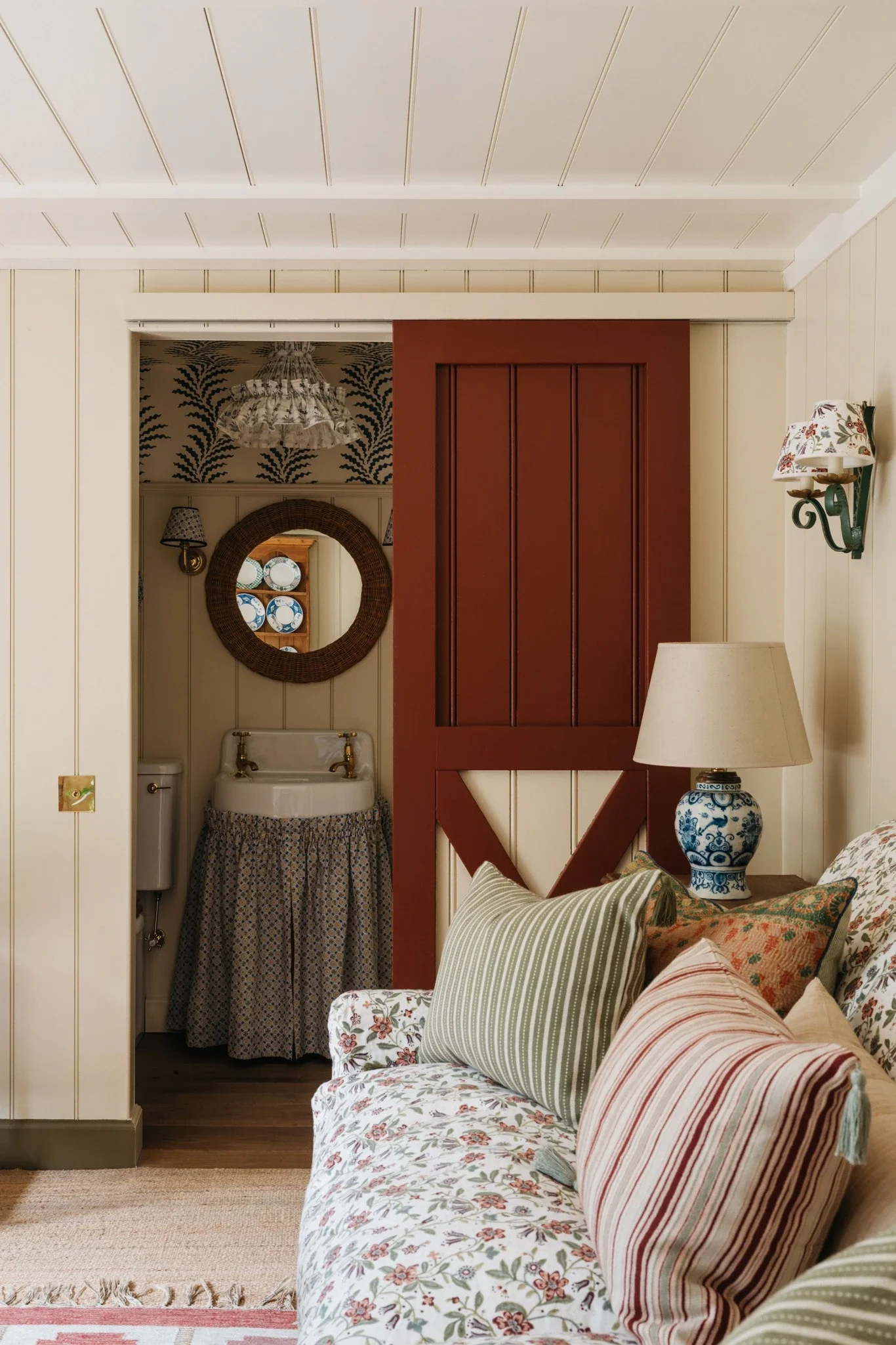 Cozy living room with floral-patterned sofa, striped pillows, a table lamp with a floral lampshade, and a doorway leading to a small room with a mirror and decorative plates.