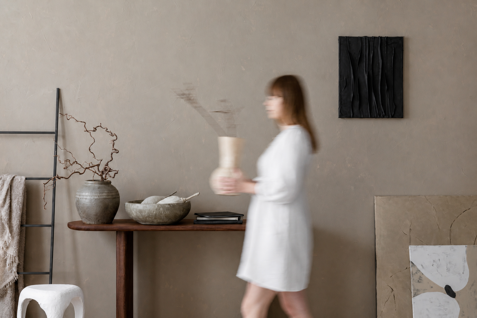 A woman in a white dress holding a ceramic vase with smoke or steam rising from it, standing in a modern, minimalistic interior with neutral-colored walls, abstract artwork, a wooden table with decorative pottery, a ladder with a blanket, and a small white stool.