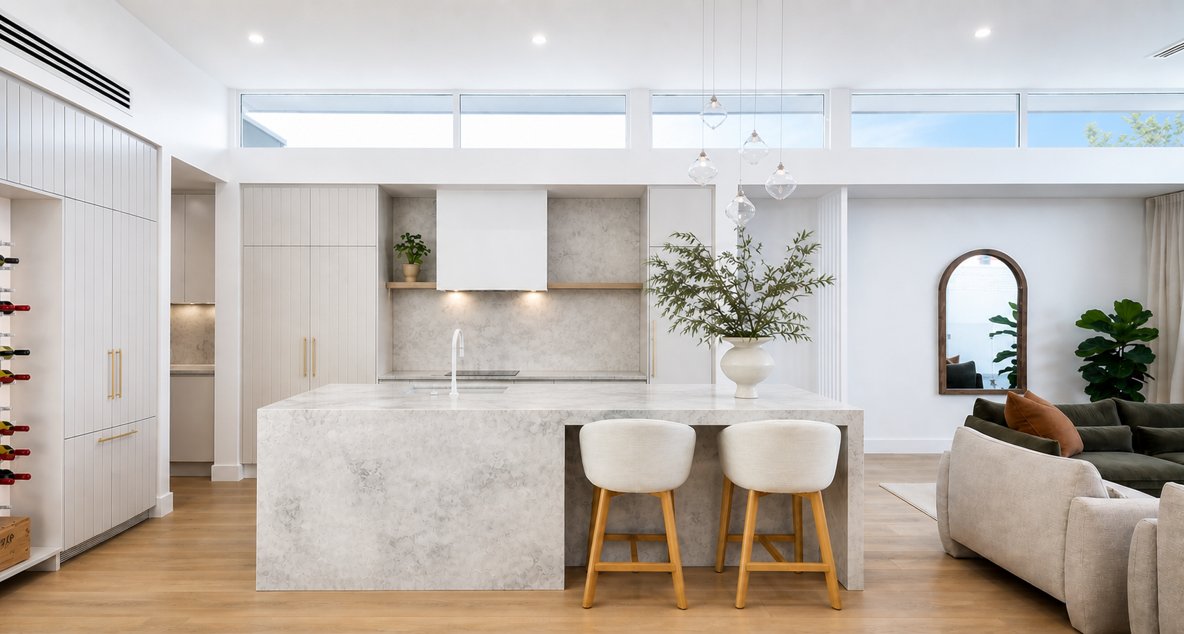 Modern open-concept kitchen and living room with white cabinetry, marble island, pendant lighting, a large potted plant, and a seating area with neutral and dark furniture.