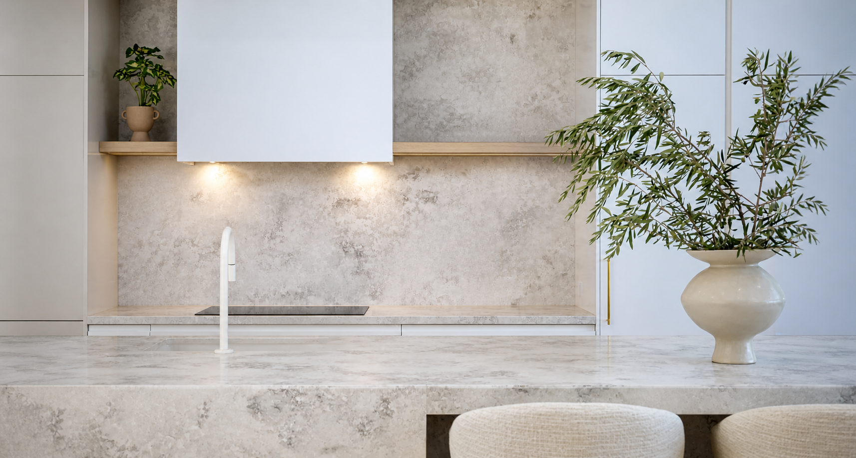 Modern minimalist kitchen with beige marble countertops, a large beige vase with green leaves, and small plants on a shelf.
