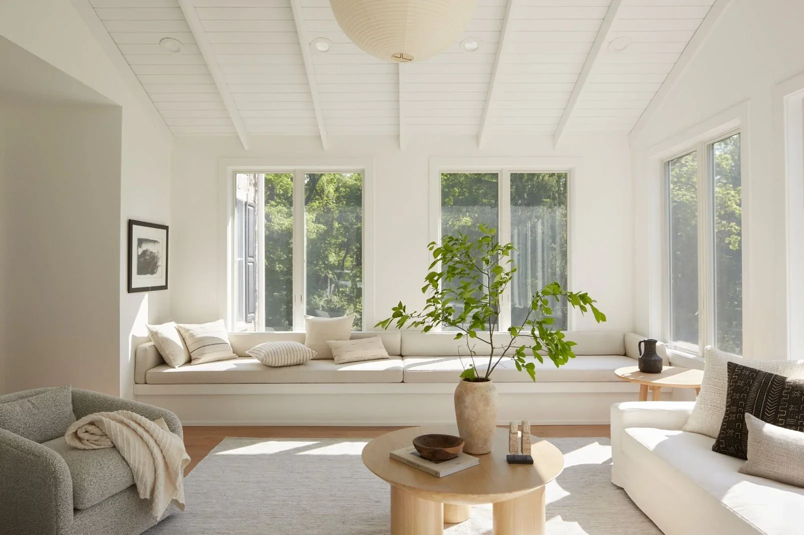 Bright living room with white walls, large windows showing greenery outside, a white built-in window seat with cushions, a central round wooden coffee table with decor, and seating including a gray armchair and a white sofa with cushions. A potted plant sits on the coffee table.