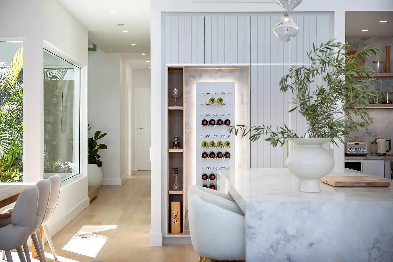 Modern kitchen with white marble counter, potted plant, and wine storage wall with wine bottles in a bright, airy space.