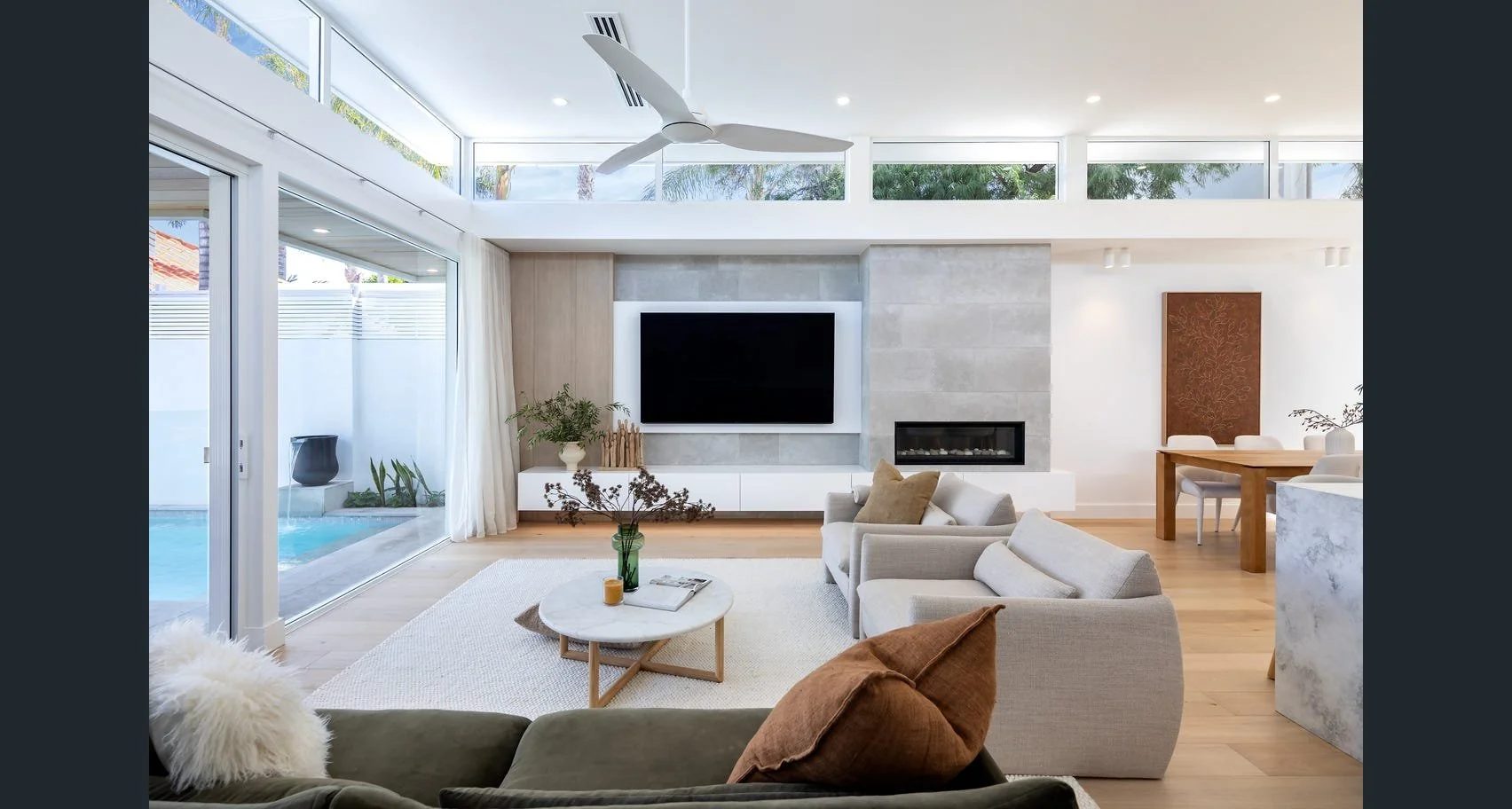 Modern living room with large sliding glass doors, a ceiling fan, and a wall-mounted television above a fireplace. There is a beige sofa with pillows, a marble coffee table, and a dining area with a wooden table and chairs. Outside, a swimming pool and greenery are visible.