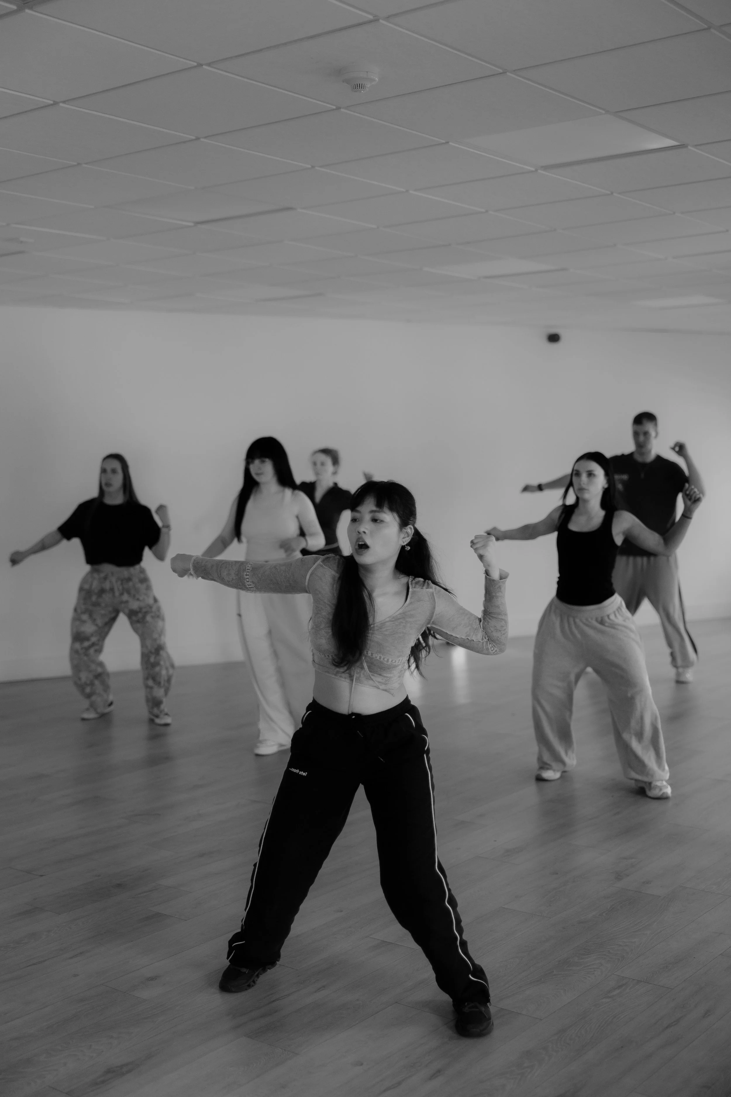 A group of people practicing dance or fitness moves in a spacious room with wooden floors and a plain white wall.