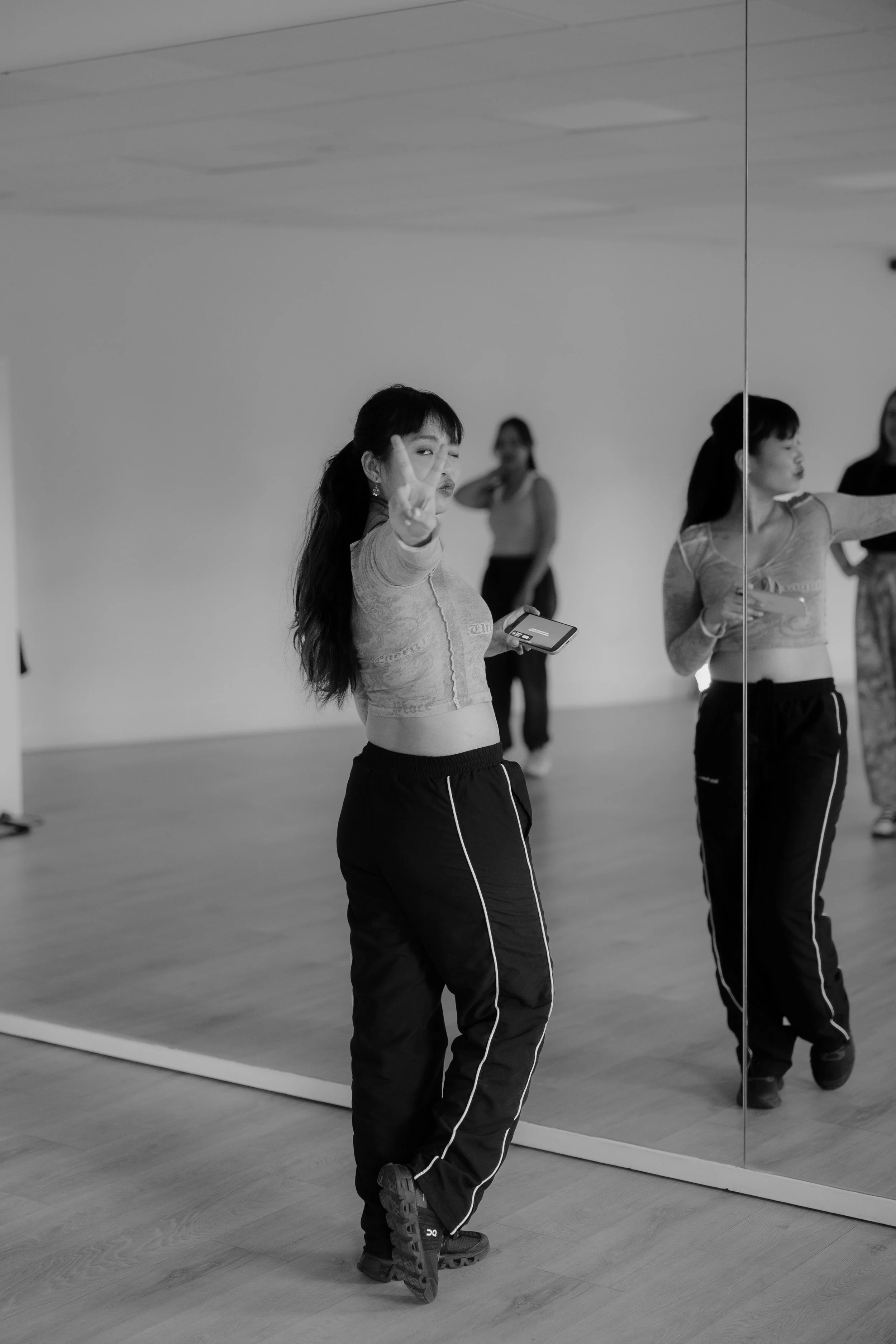A woman dressed in workout clothes standing in front of a large mirror in a dance or fitness studio, making a peace sign with her hand and holding a smartphone in her other hand. Other women are visible in the background.