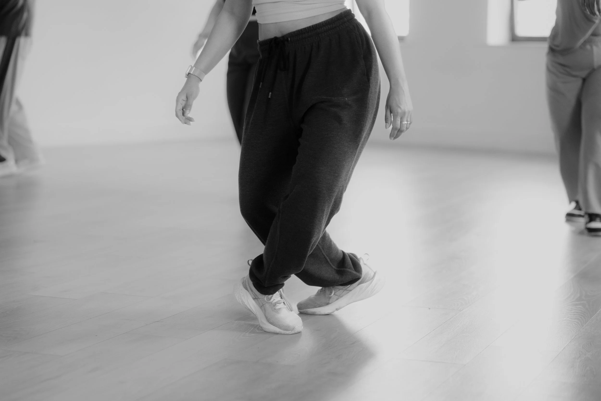 People practicing dance or movement in a studio, focusing on their legs and feet, with a wooden floor and natural light.