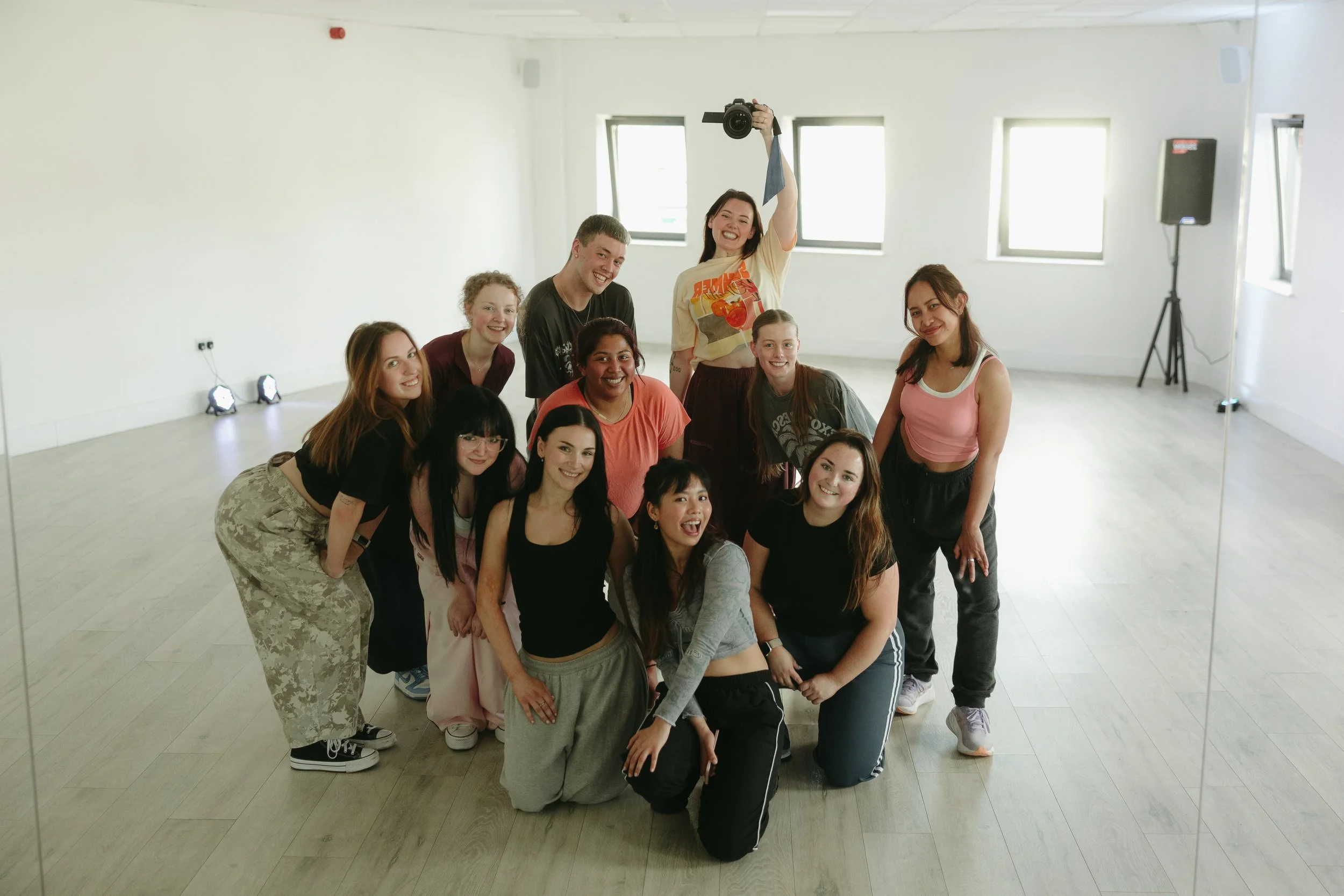 A group of young women and men posing together in a well-lit room with large windows. One woman is holding a camera above her head, smiling. The group appears happy and energetic, dressed casually, in a dance or fitness studio with light-colored floors and walls.