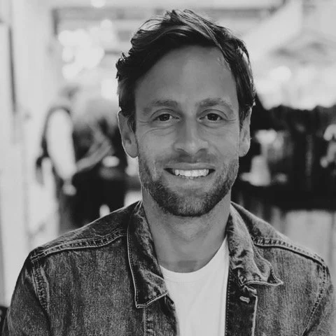 Black and white photo of a smiling man with short dark hair, light beard, wearing a denim jacket and white shirt indoors.