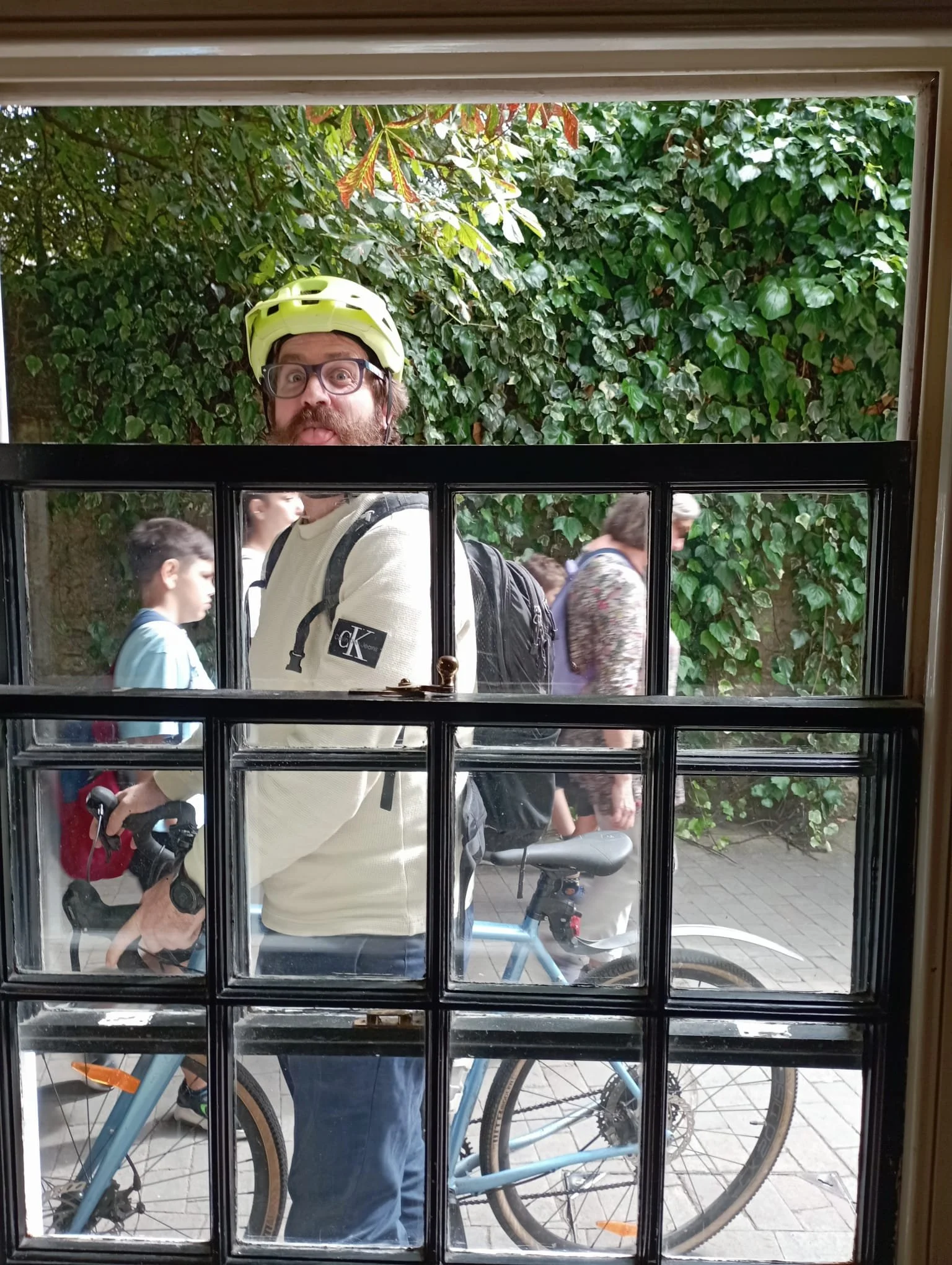 Person with a beard and glasses wearing a yellow helmet looking through a window, with multiple people and bicycles outside against a background of green leafy bushes.