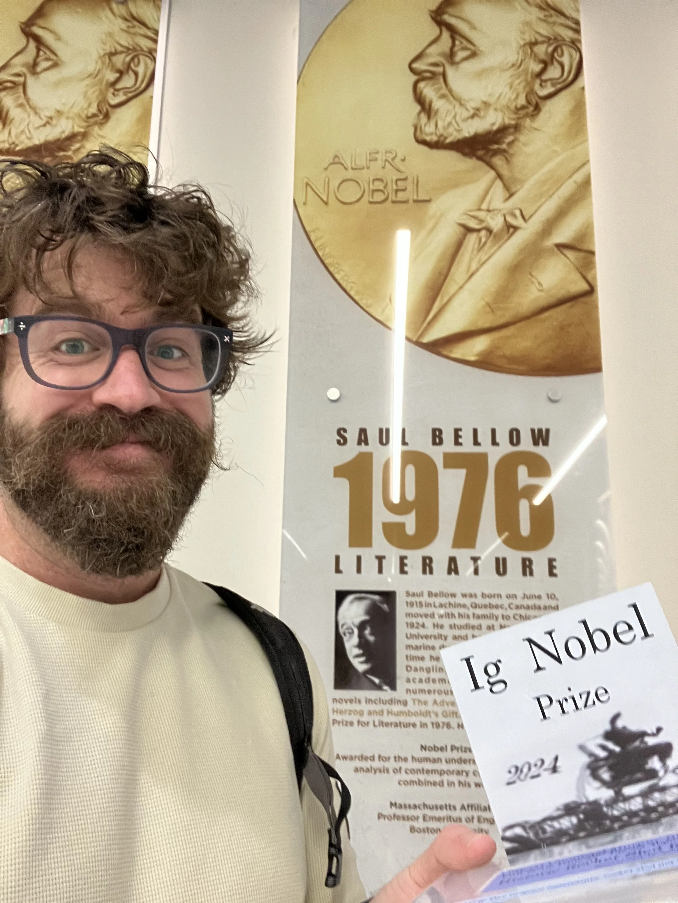 Saul Newman with glasses, a beard, and curly hair holding an Ig Nobel prize in front of the display honoring Nobel laureate Saul Bellow.