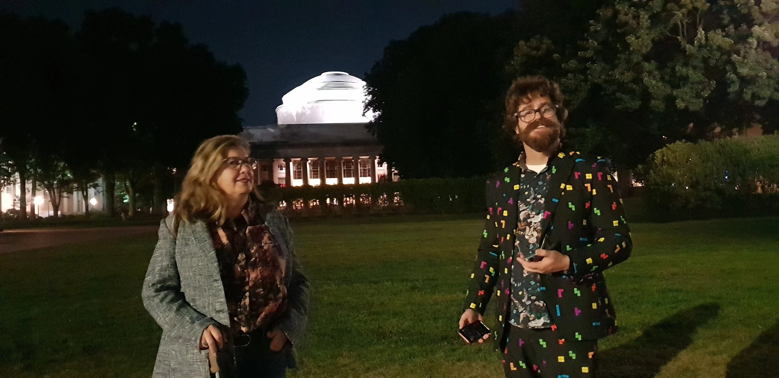 Two people, a woman on the left and a man on the right, standing outdoors at night with a lit building in the background. The woman has long wavy blonde hair, glasses, and is wearing a grey blazer with a floral shirt. The man has curly hair, a beard, glasses, and is wearing a colorful, patterned blazer and shirt, holding a phone in his right hand.