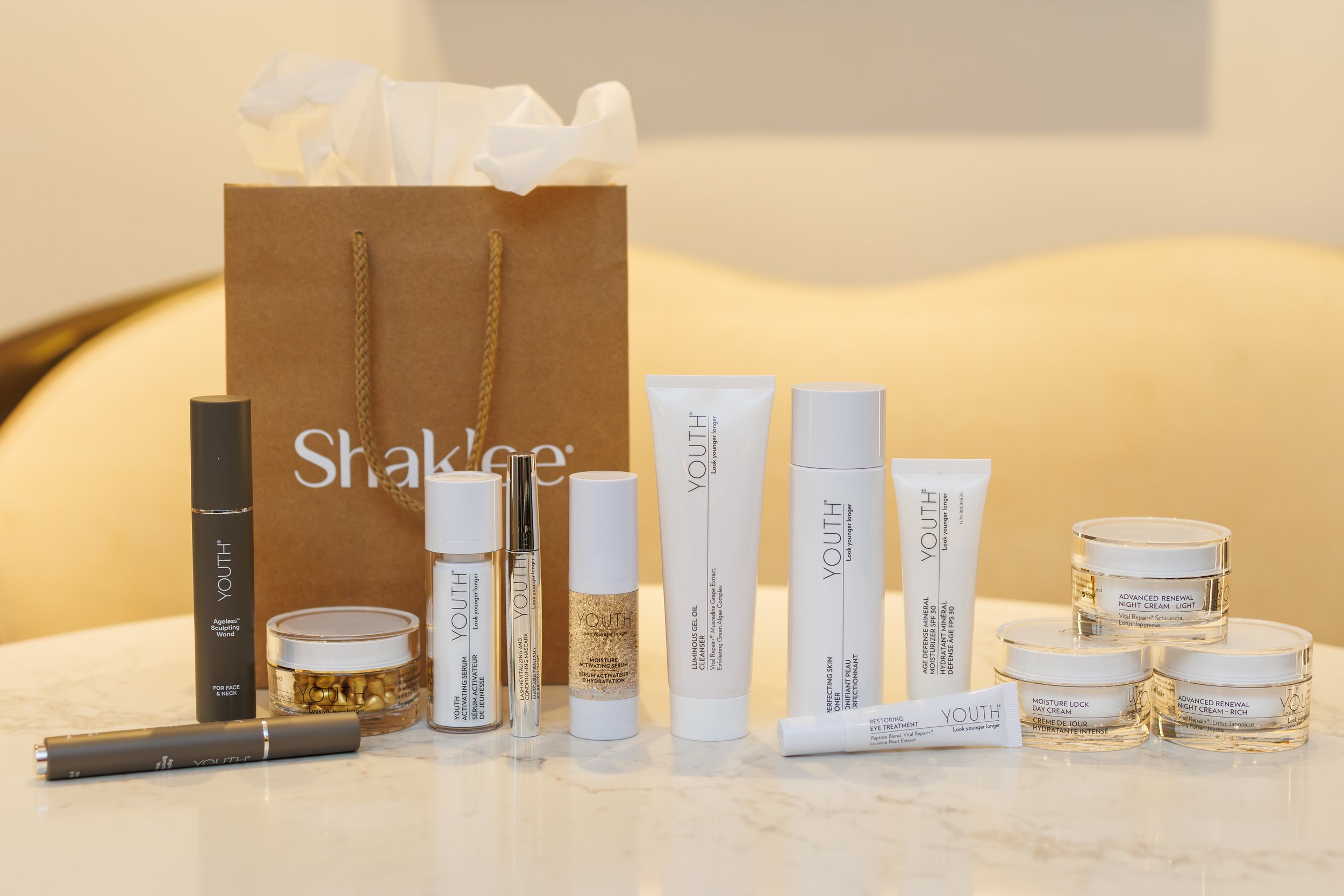 A collection of skincare and beauty products, including creams, serums, and a masking wand, arranged on a white surface in front of a brown shopping bag labeled 'shake' and a few small jars of night cream.