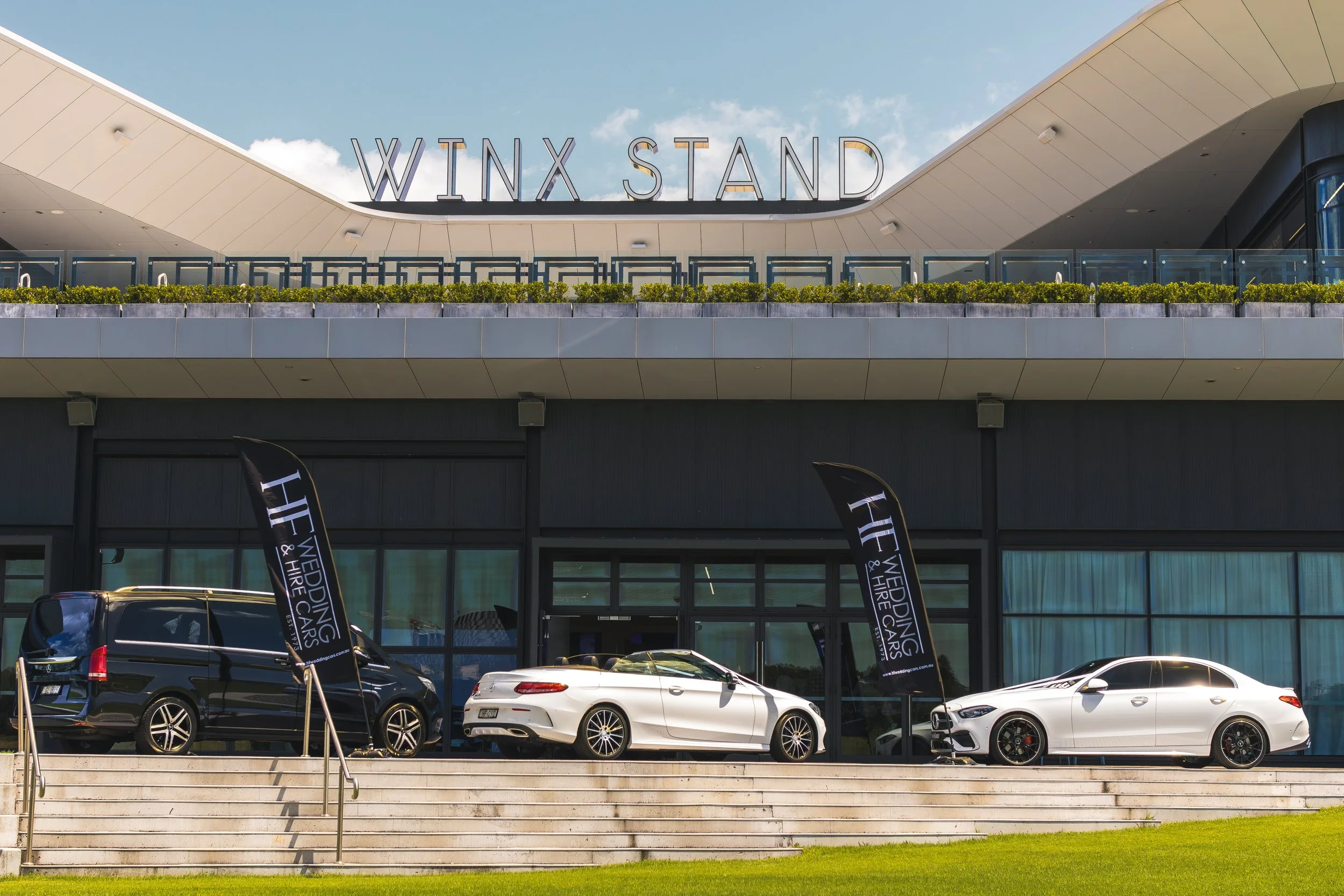 Exterior view of Winx Stand at a car dealership with several new cars parked in front and flags displaying the dealership's name.