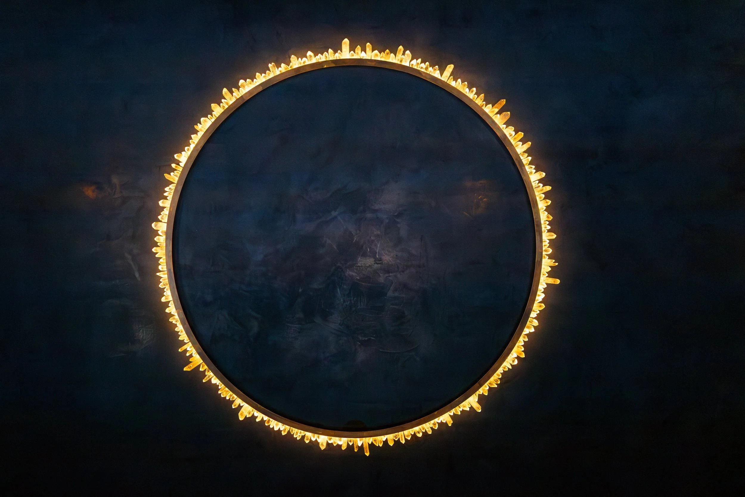 Half of a round mirror with a yellowish crystal light border, mounted on a dark wall.