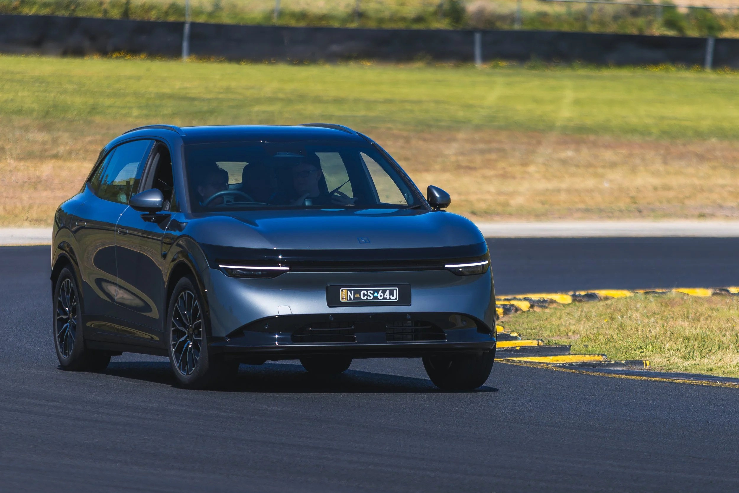 A modern black electric car driving on a paved road with a grassy area and a black fence in the background.