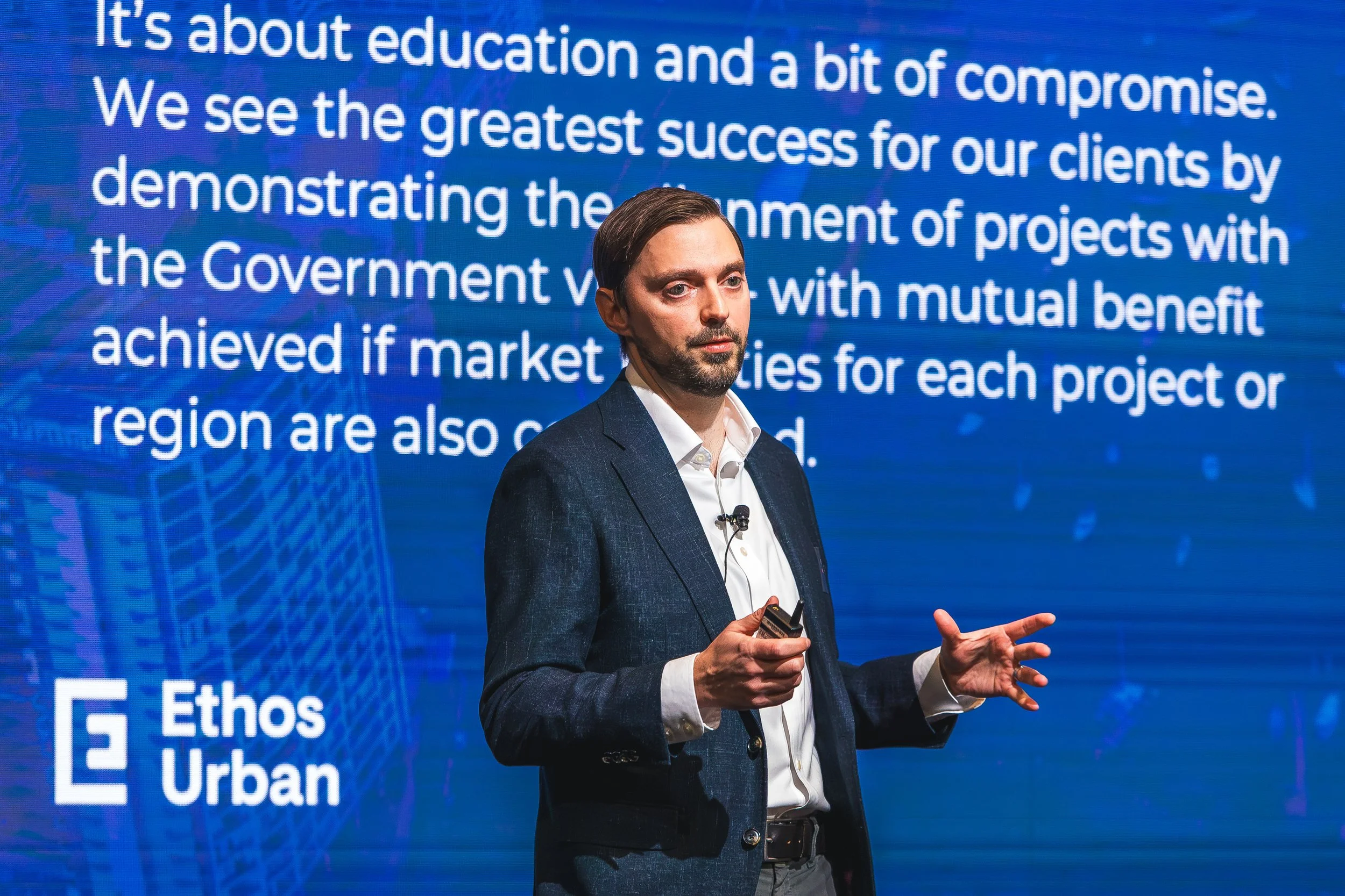 A man in a dark suit and white shirt giving a presentation in front of a blue screen with text. He holds a clicker and gestures with his right hand.