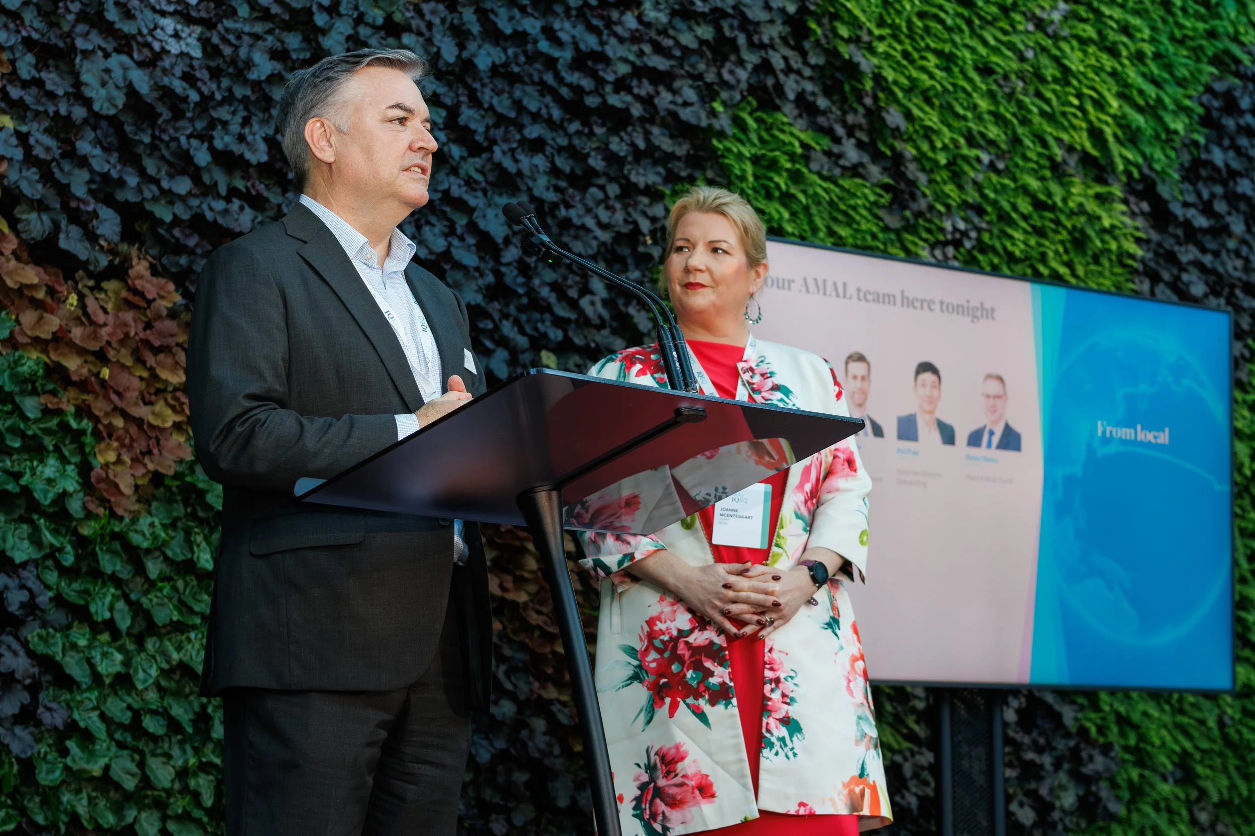 A man in a dark suit speaking at a podium with a woman in a floral blazer standing beside him, both on a stage with a colorful leafy background and a banner.
