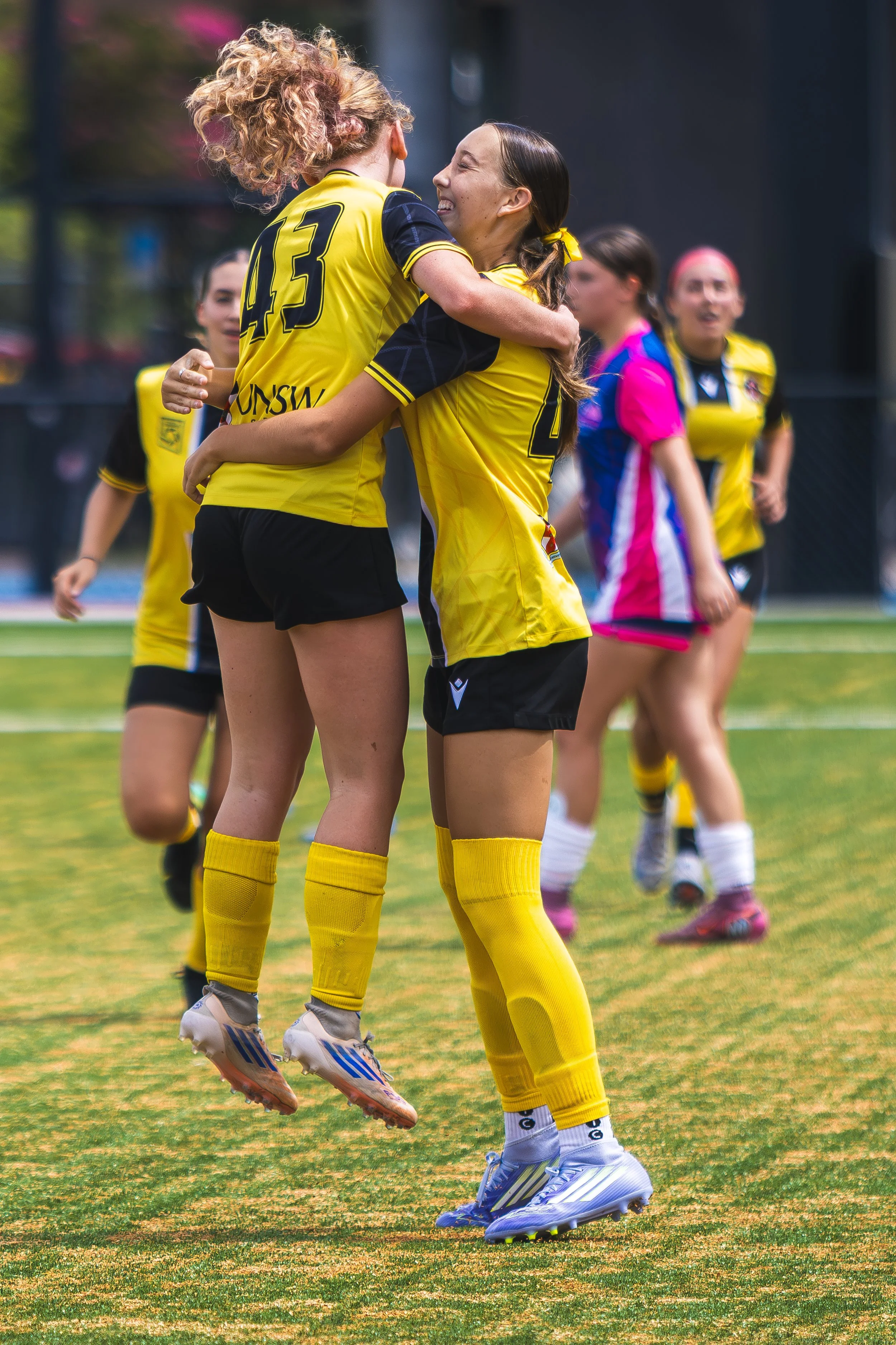 Two female soccer players in yellow uniforms are celebrating by hugging on the field, with other players and a chain-link fence in the background.
