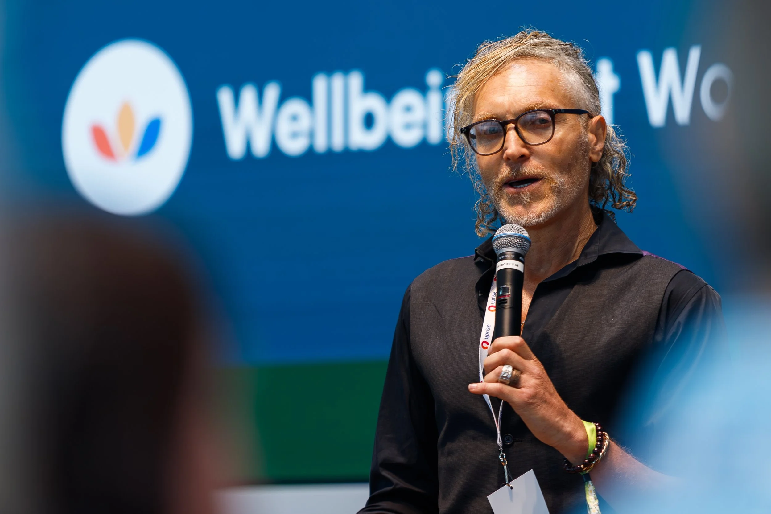 A man with glasses and gray hair speaking into a microphone at an indoor event with a blue background that has the word 'Wellbeing' and a colorful logo.