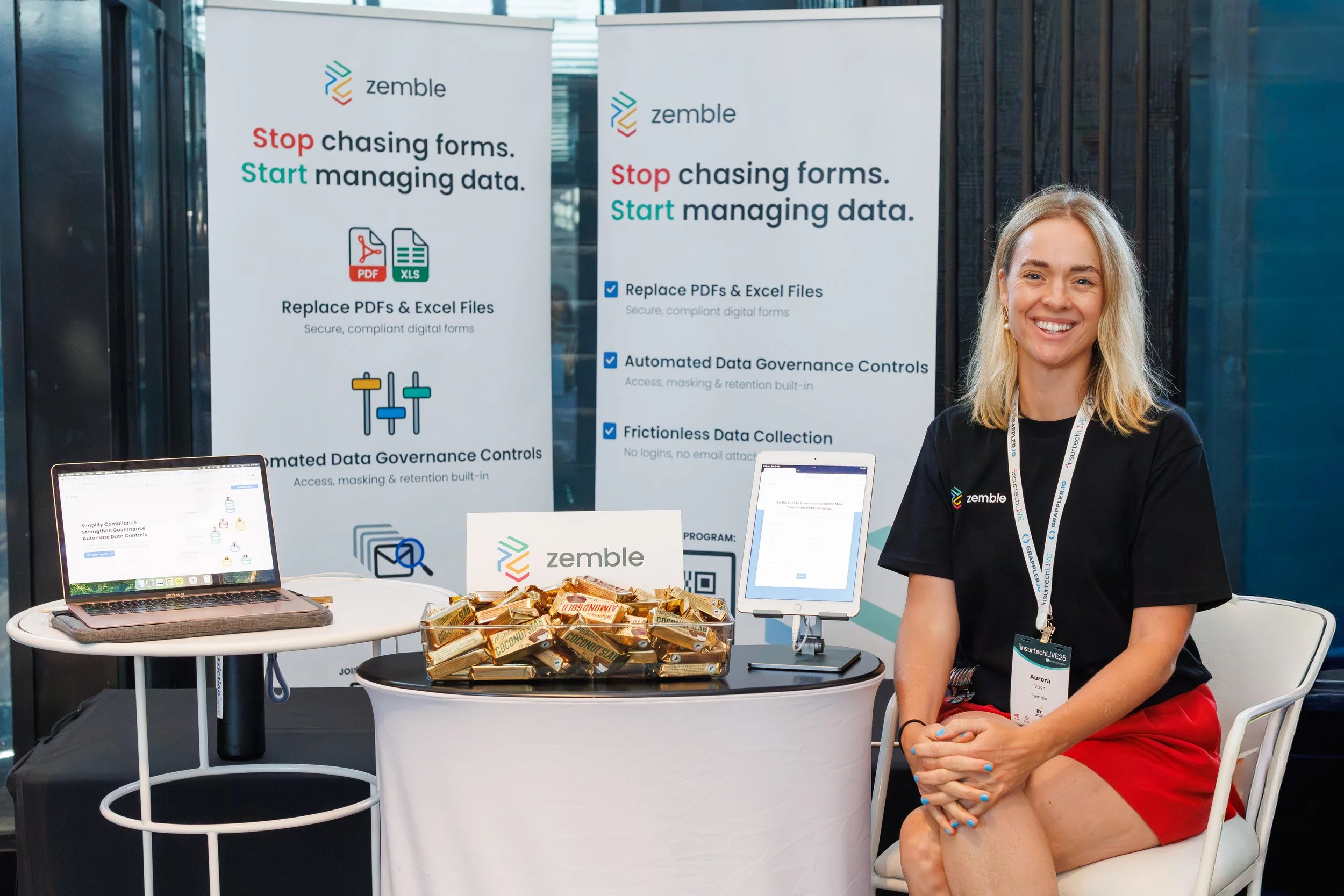 A woman with blonde hair smiling at a technology trade show booth. The booth features a Zumble banner promoting digital forms and data management tools, with a laptop, tablets, and snacks on the table in front of her.