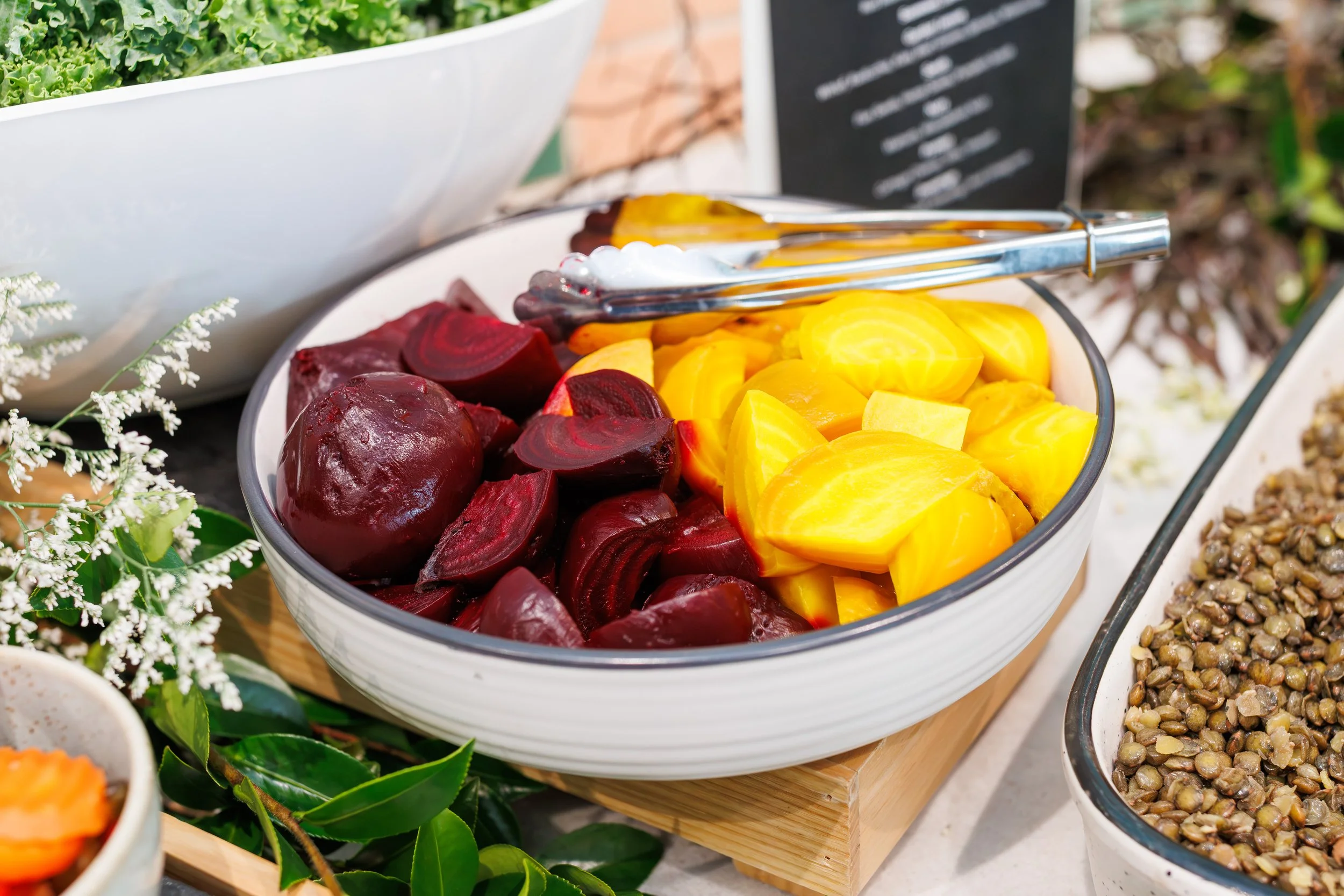 Bowl of sliced beets and yellow squash or pumpkin at a buffet or salad bar. Tongs rest on top of the bowl.