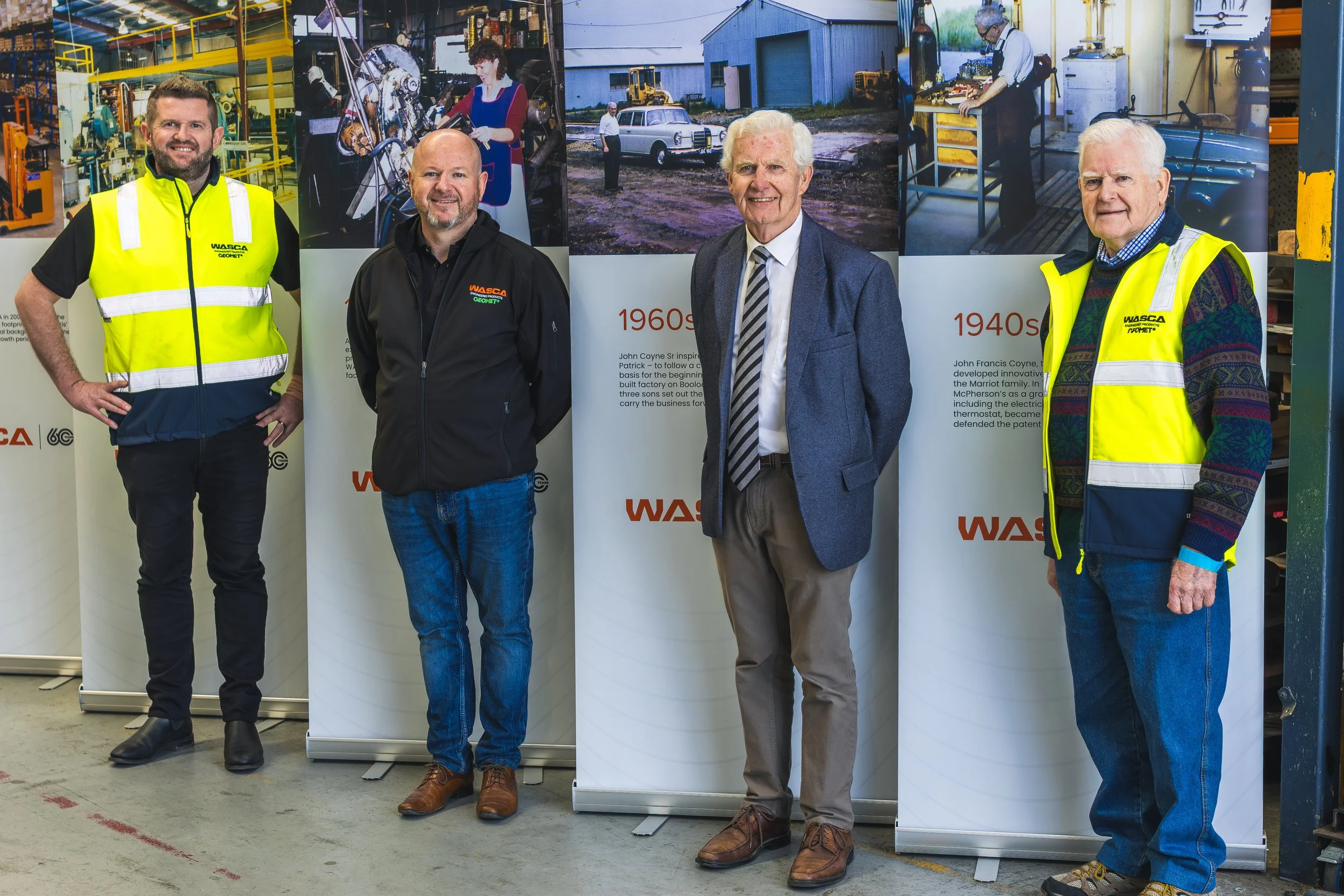 Four men standing in front of informational posters about the history of a company, in an industrial setting. The posters depict photos and text about the company's development from the 1940s and 1960s.