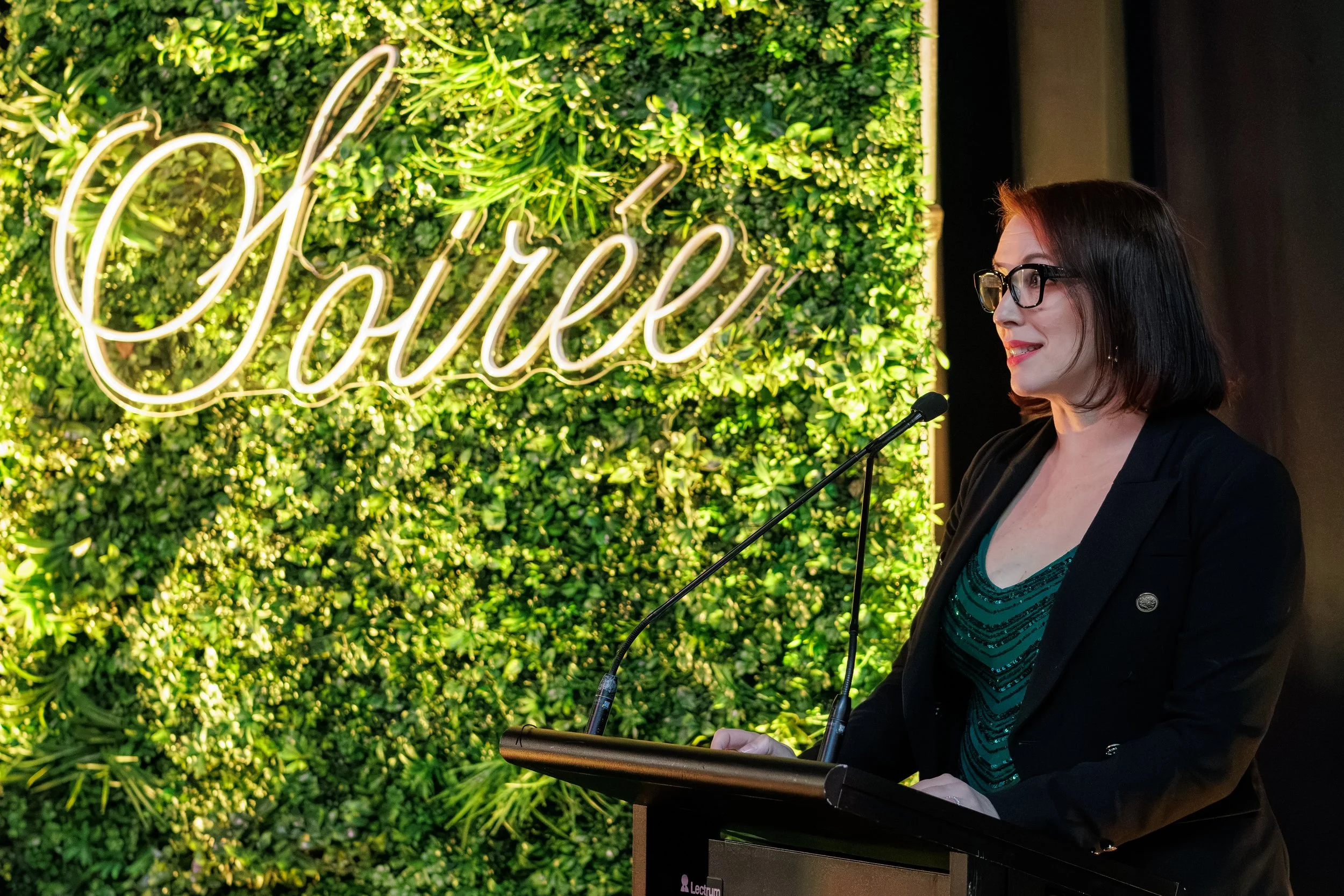 A woman in glasses, wearing a dark blazer and teal top, stands at a podium with a microphone in front of a greenery wall with a neon sign spelling 'Charmed' in cursive.