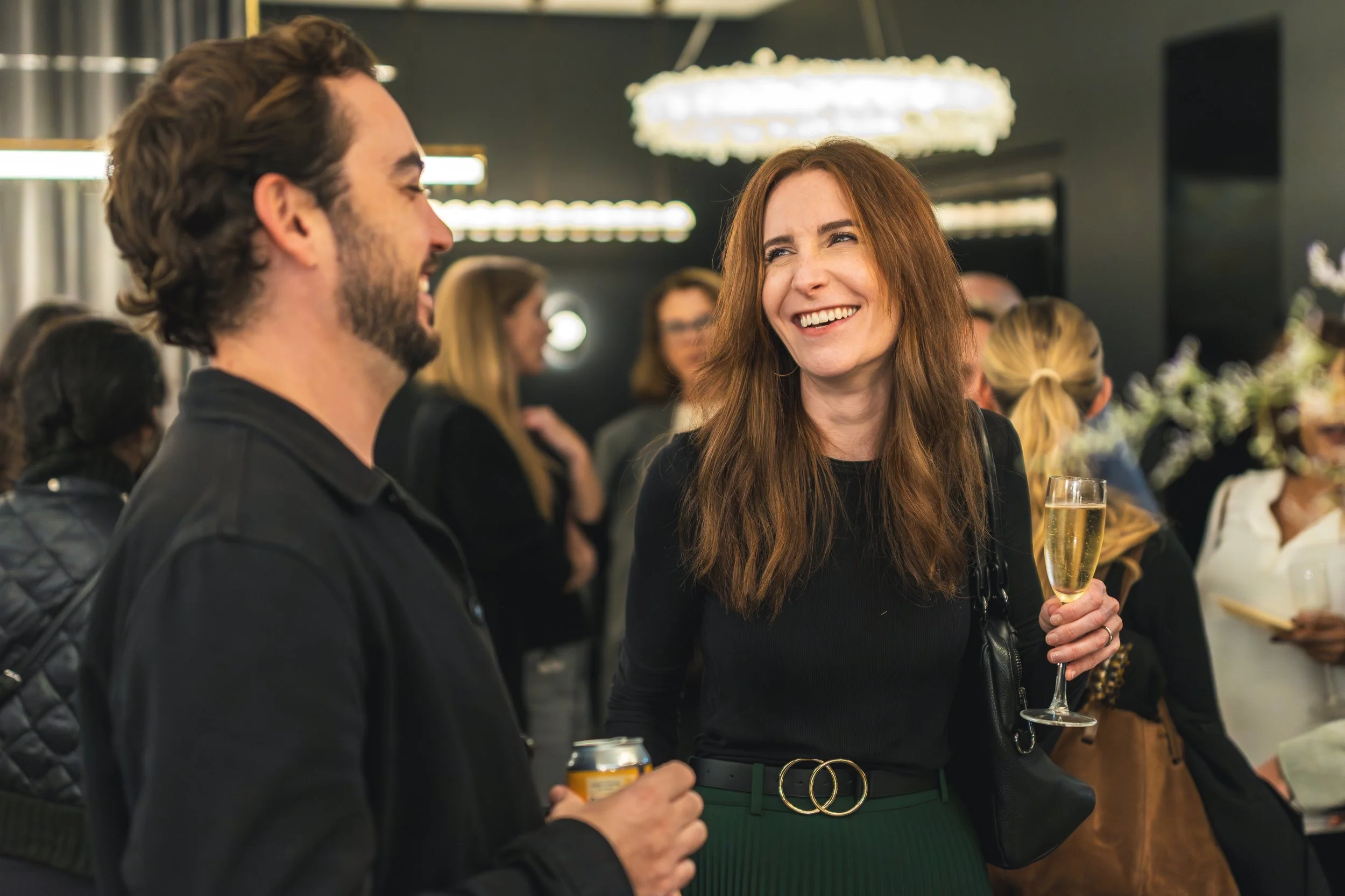 Two people, a man and a woman, are smiling and socializing at a party. The woman is holding a champagne flute and a woman in the background is holding a glass of champagne. The setting appears to be a lively indoor event with warm lighting and multip