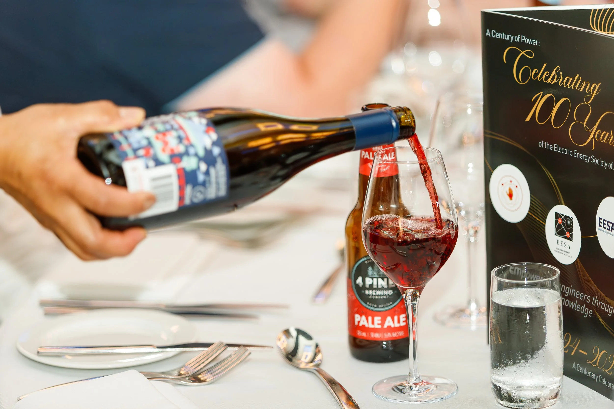 Person pouring red beer into a wine glass at a celebration event, with a sign in the background that reads "Celebrating 100 Years of the Electric Energy Society."
