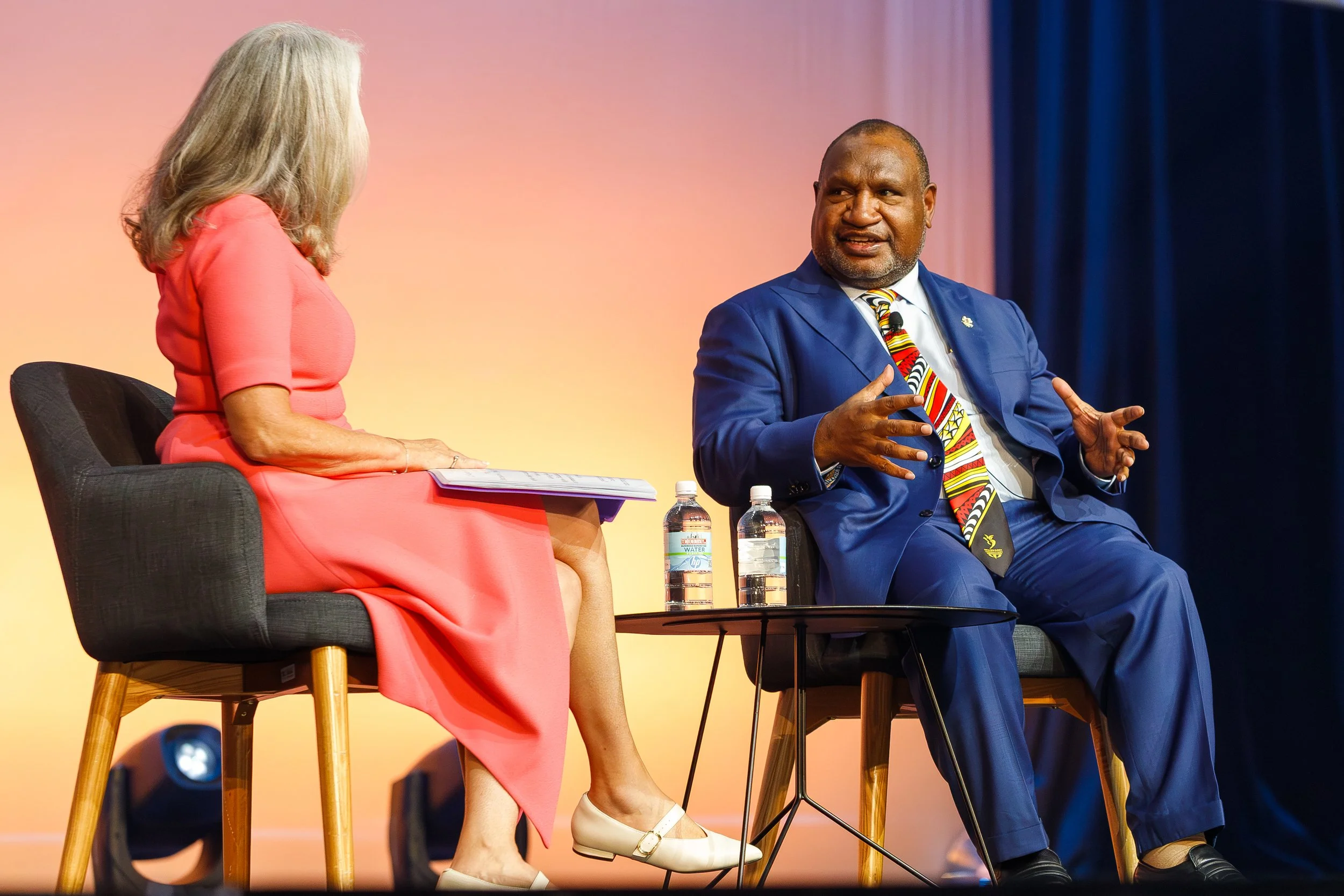 A woman in a pink dress and beige heels and a man in a blue suit and striped tie are sitting on stage and talking to each other. The woman is facing away from the camera, and the man is facing toward her.
