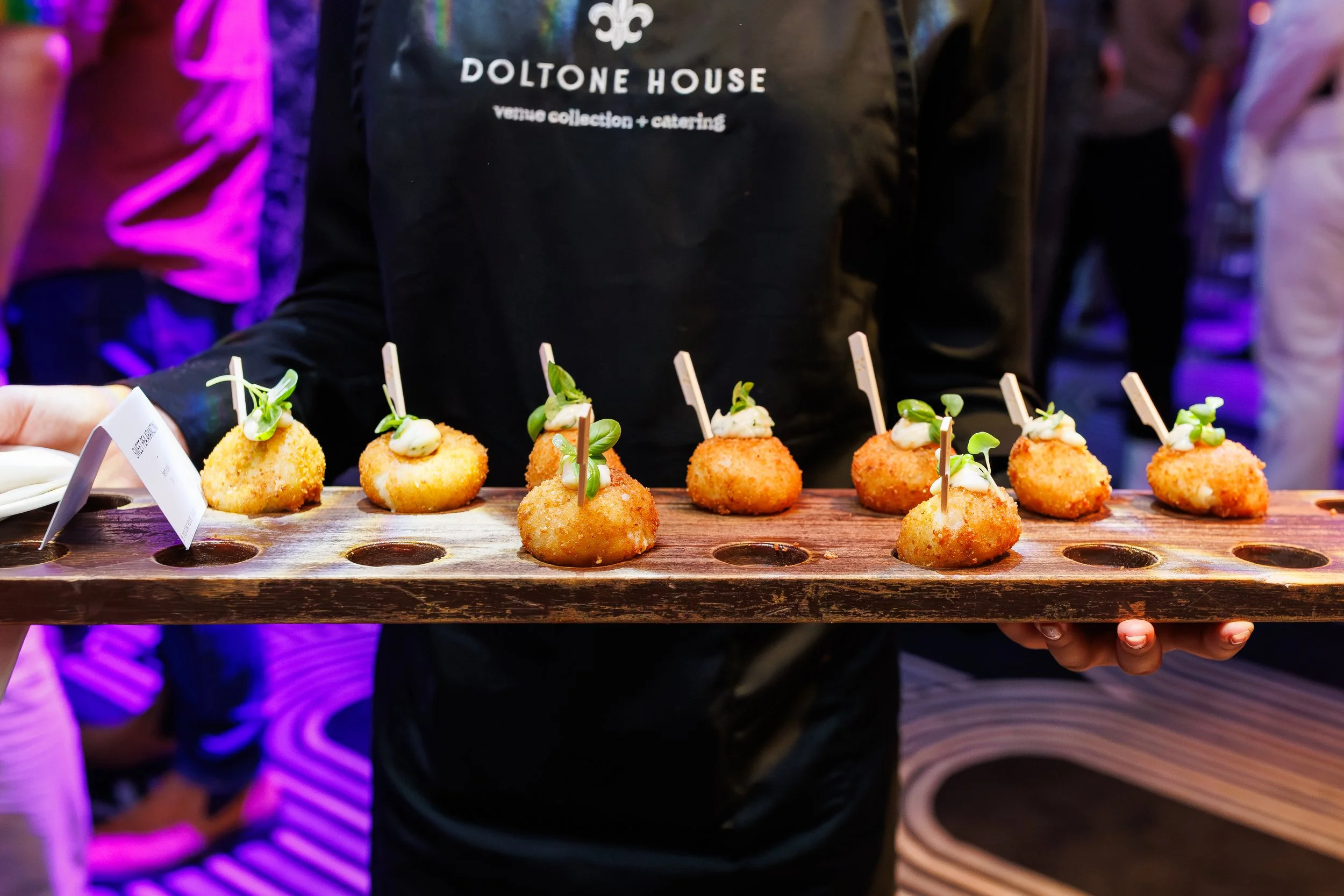 A person holding a wooden tray with several golden-brown fried appetizers topped with white sauce and small green herbs, set against a blurred background with colorful lighting.