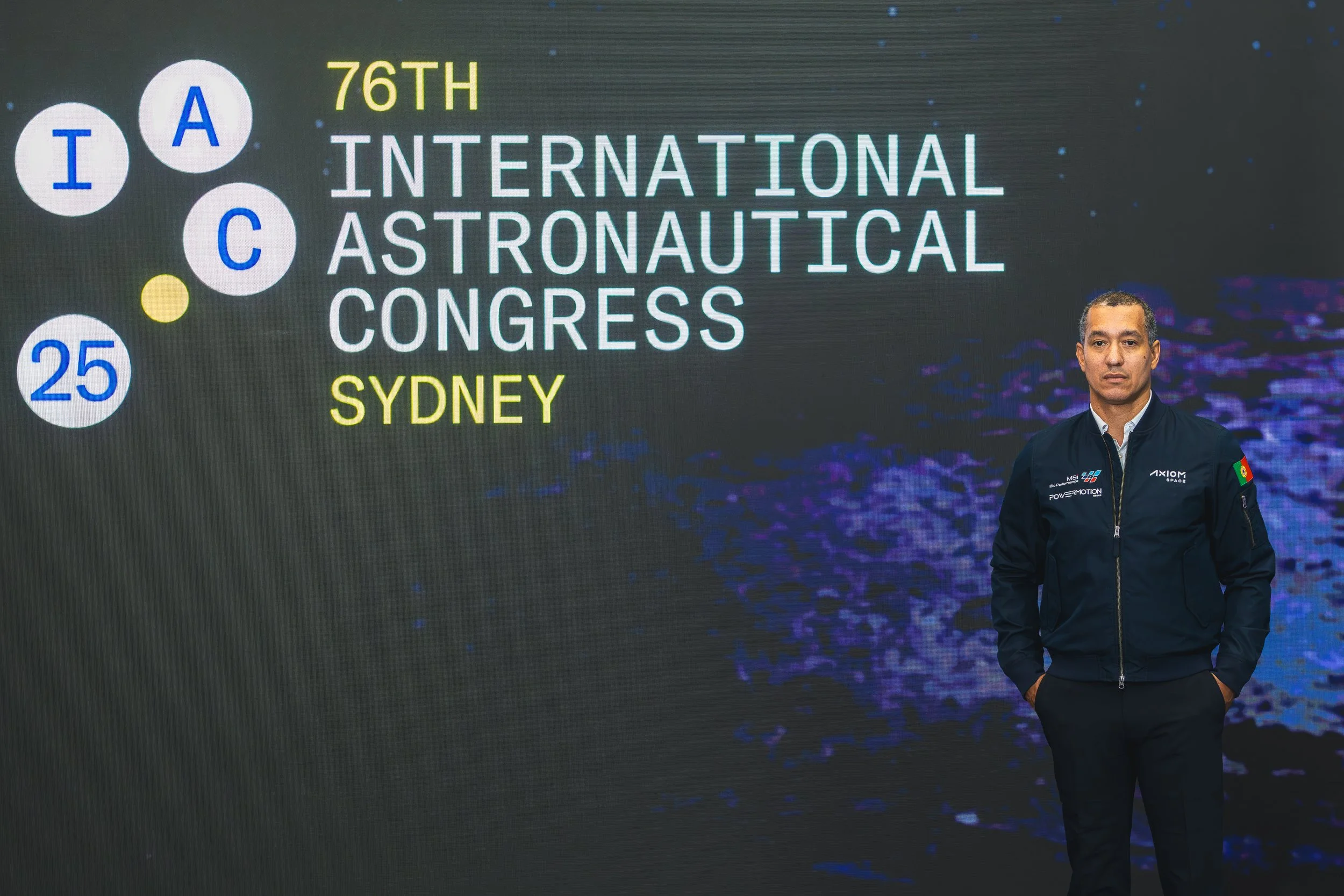 A man in a dark jacket with a Portuguese flag patch on the sleeve standing in front of a large display screen. The screen shows the text '76th International Astronautical Congress Sydney' with the words 'International' and 'Congress' in large white l