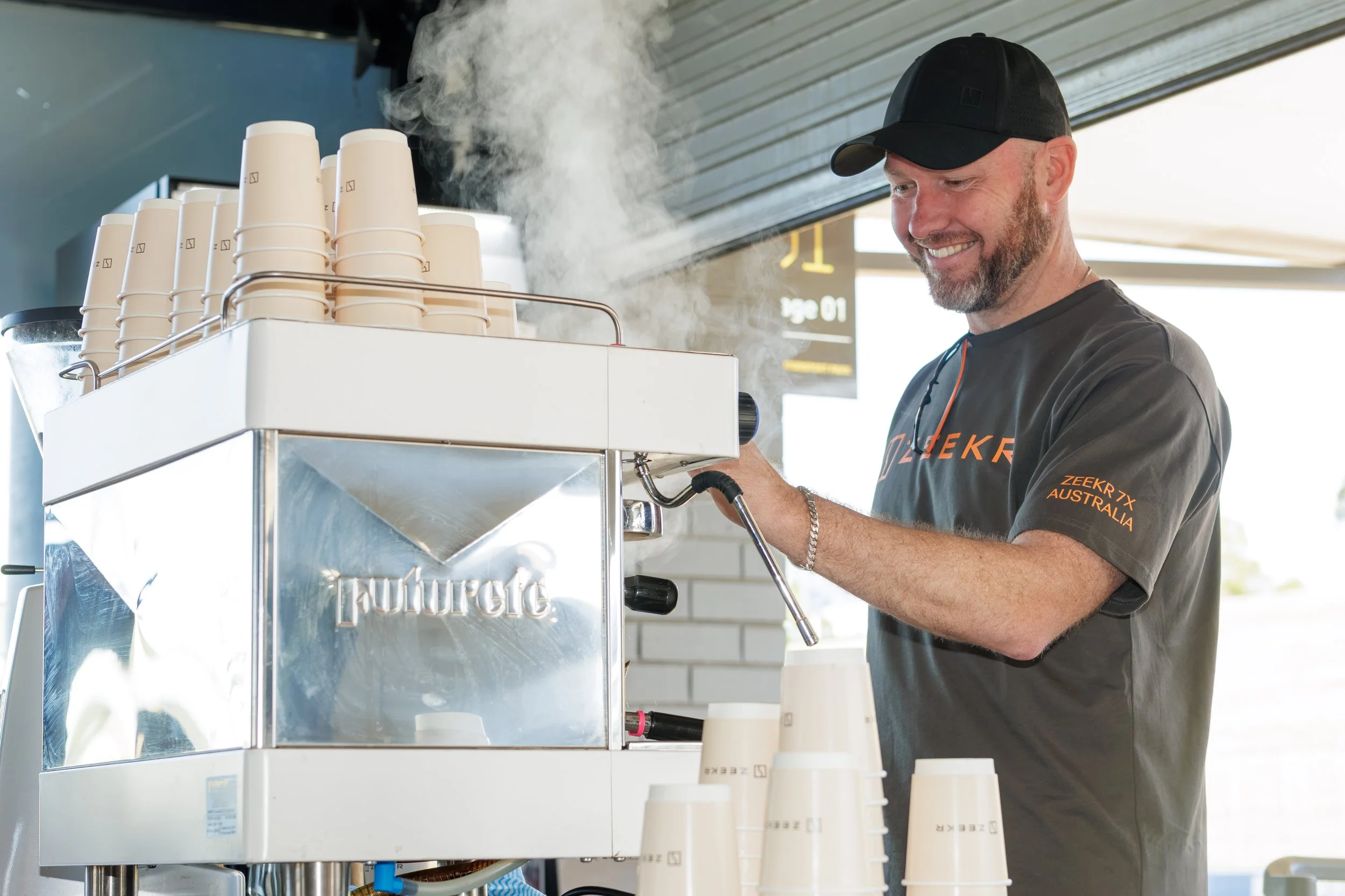 A man with a beard and a black cap making coffee with a professional espresso machine, with stacked disposable cups nearby.