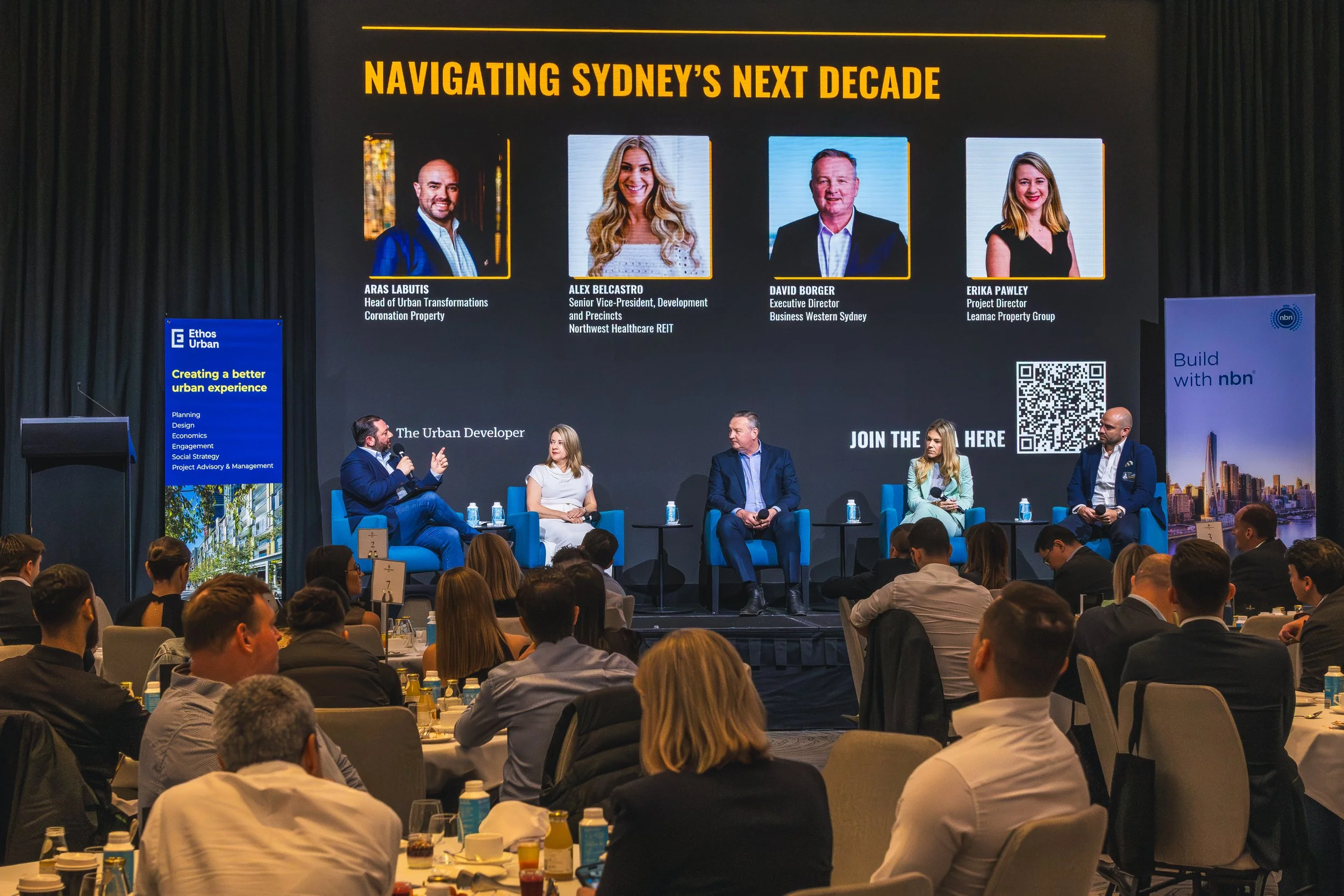 A large conference room with a stage, where five panelists are seated facing the audience. A large screen behind them displays the title 'Navigating Sydney's Next Decade' with photos and titles of four speakers: Aras Labutis, Alex Belcastro, David Bo