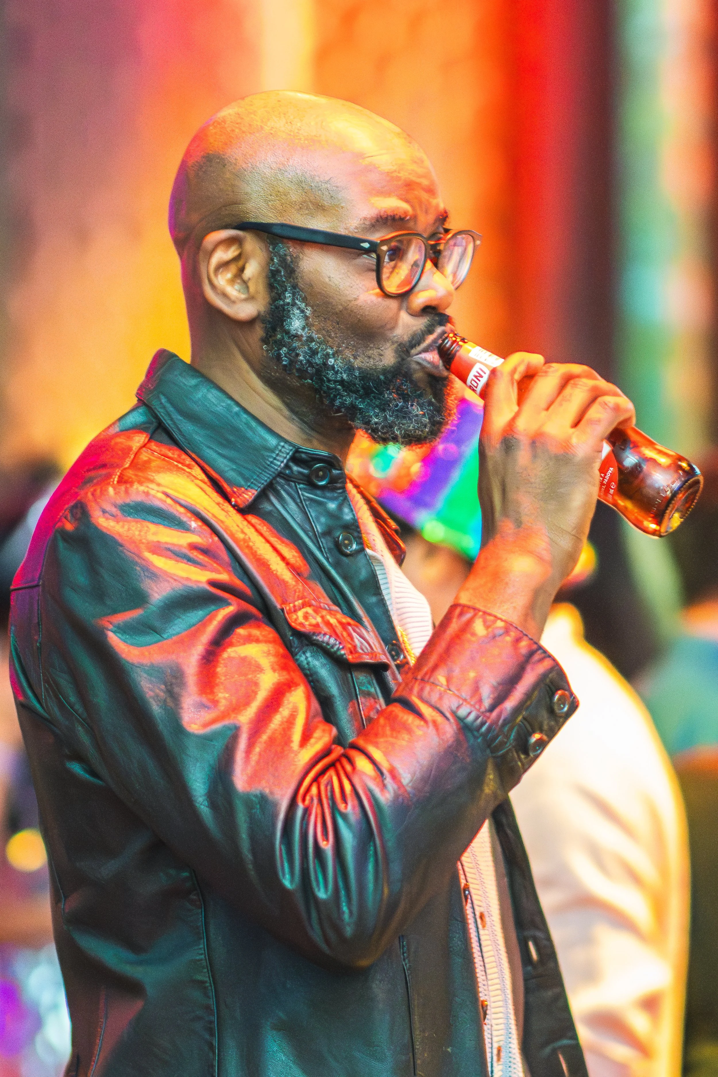 A man with glasses and a beard, dressed in a colorful, shiny leather jacket, drinking from a small glass bottle with a red and white label, in a setting with colorful, blurred lights.