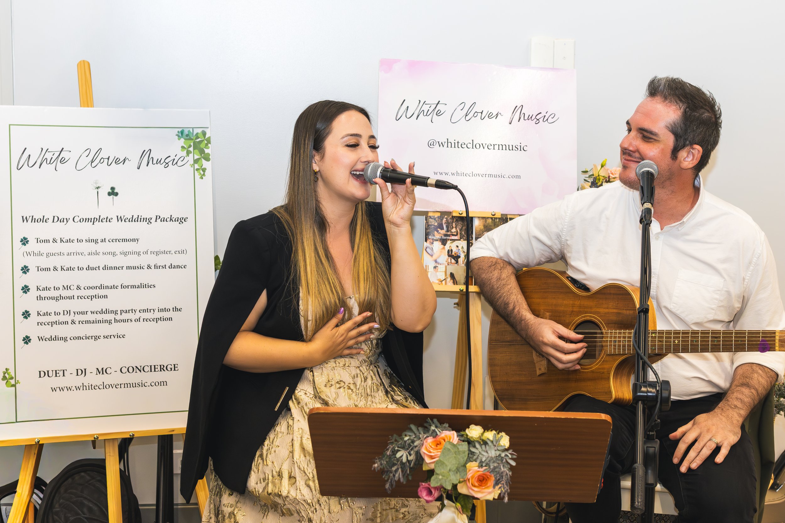 A woman singing into a microphone while a man plays a guitar beside her. They are performing at an event with signs indicating "White Clover Music" and details about wedding entertainment services.