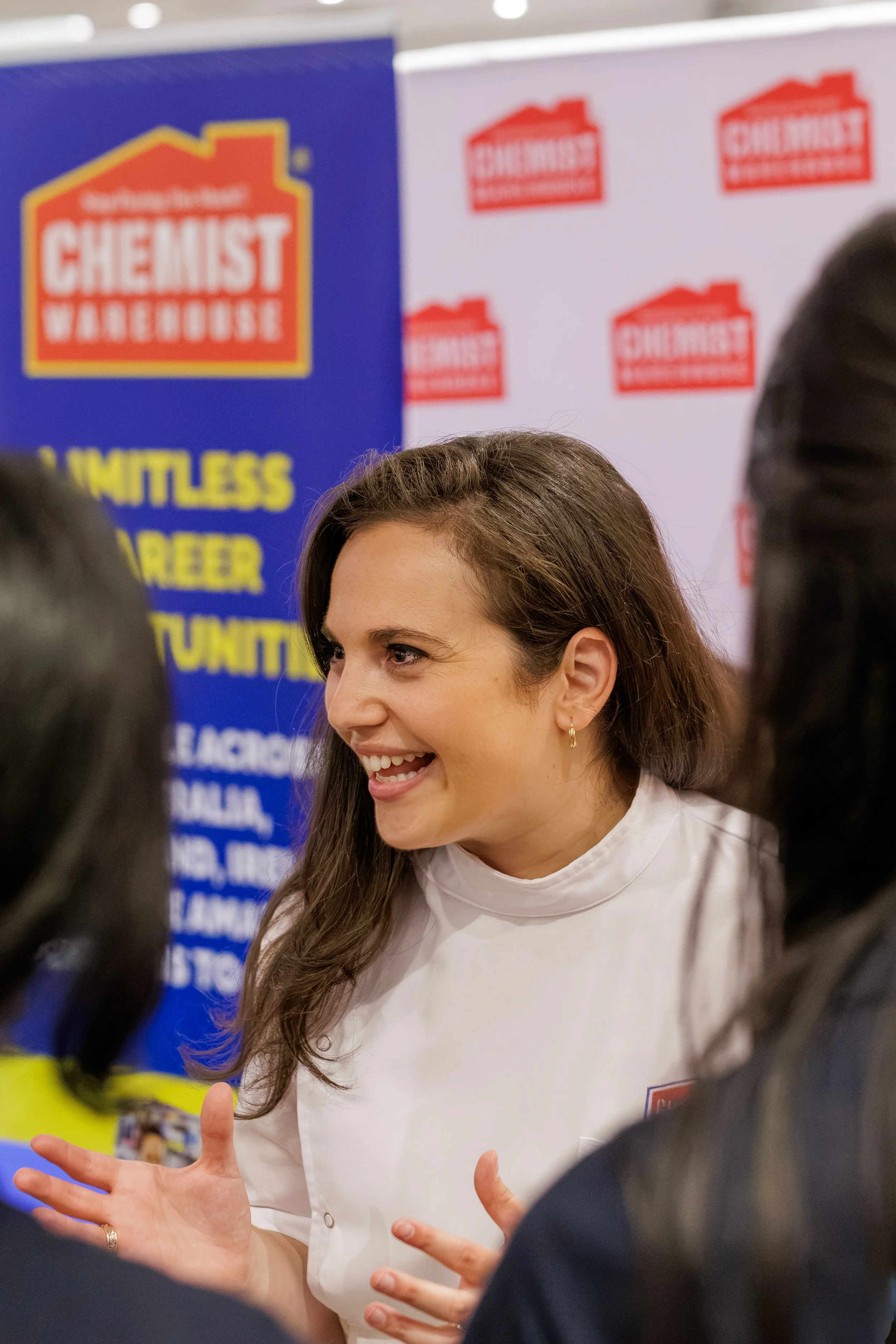 A woman with long brown hair smiling and gesturing with her hands during a discussion at a political event, with banners in the background displaying the names 'CHEAT' and 'WARS'.